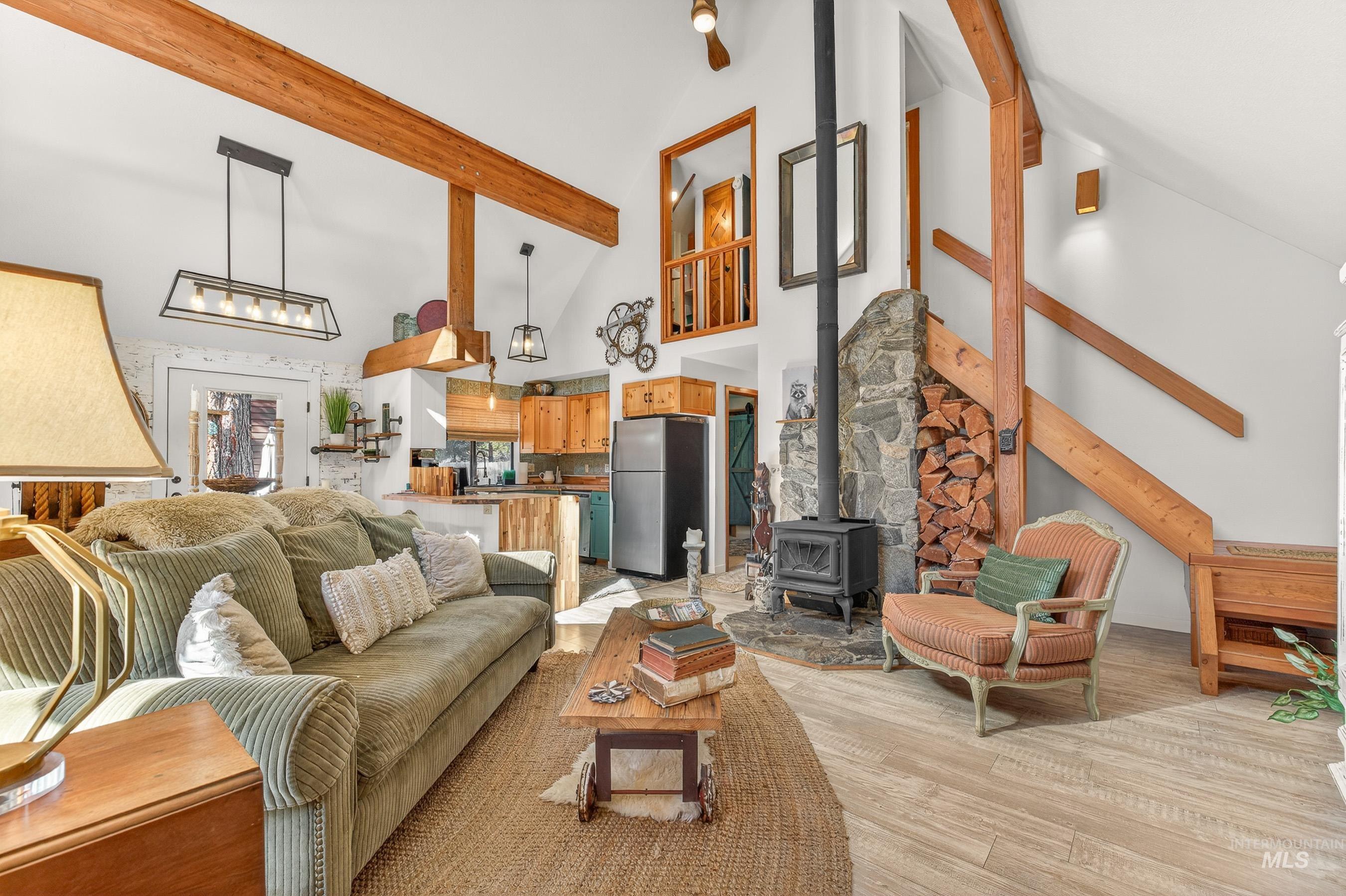 Vaulted ceiling with exposed wood beams, stone-faced wood-burning stove, light wood-finish flooring, and built-in wood storage niche