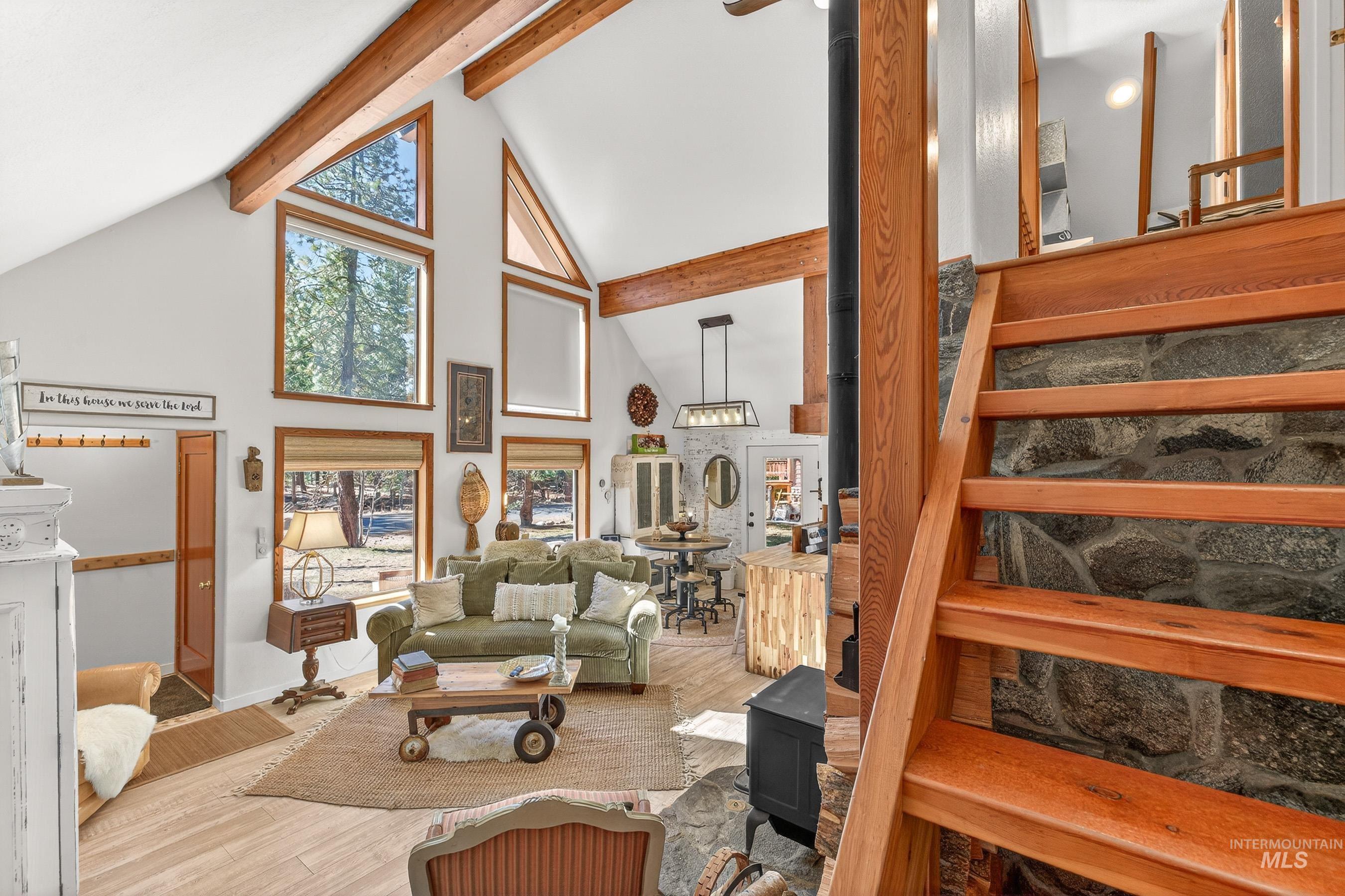 Vaulted living area featuring exposed wood beams, high ceilings, large windows, a stone-faced fireplace with a wood-burning stove, and wood-finish flooring