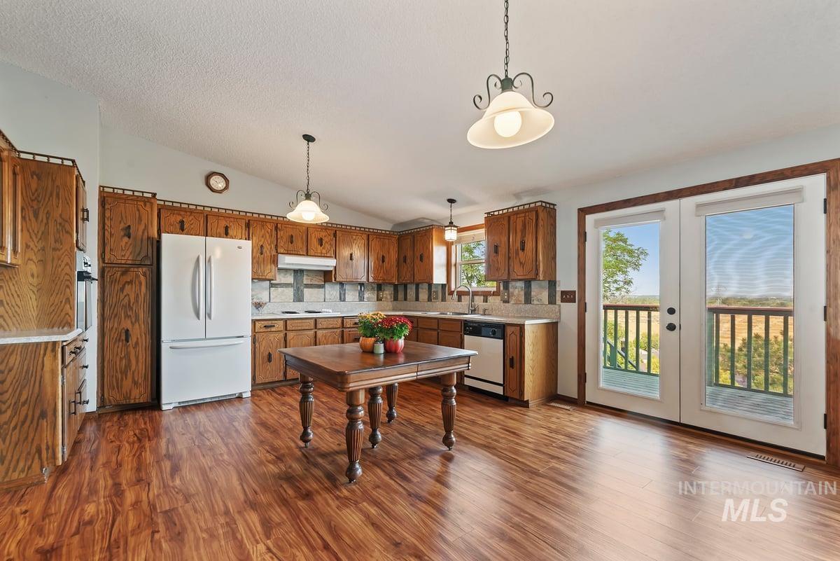 Kitchen with lofted ceiling, brown cabinetry, dark wood-type flooring, white appliances, and hanging light fixtures