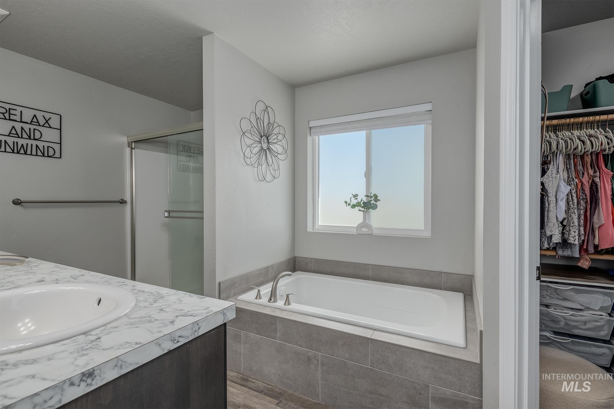 Bathroom featuring a built-in soaking tub with tile surround, a single vanity with a white countertop and integrated sink, a glass-enclosed shower, and a dedicated closet space with built-in shelving and hanging rods