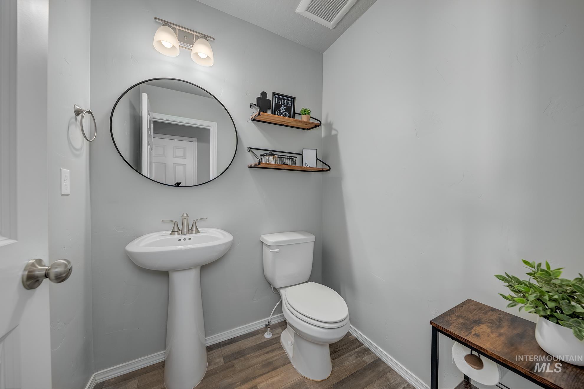 Pedestal sink with a circular mirror, brushed nickel light fixture, toilet, wood-finish flooring, and light gray walls