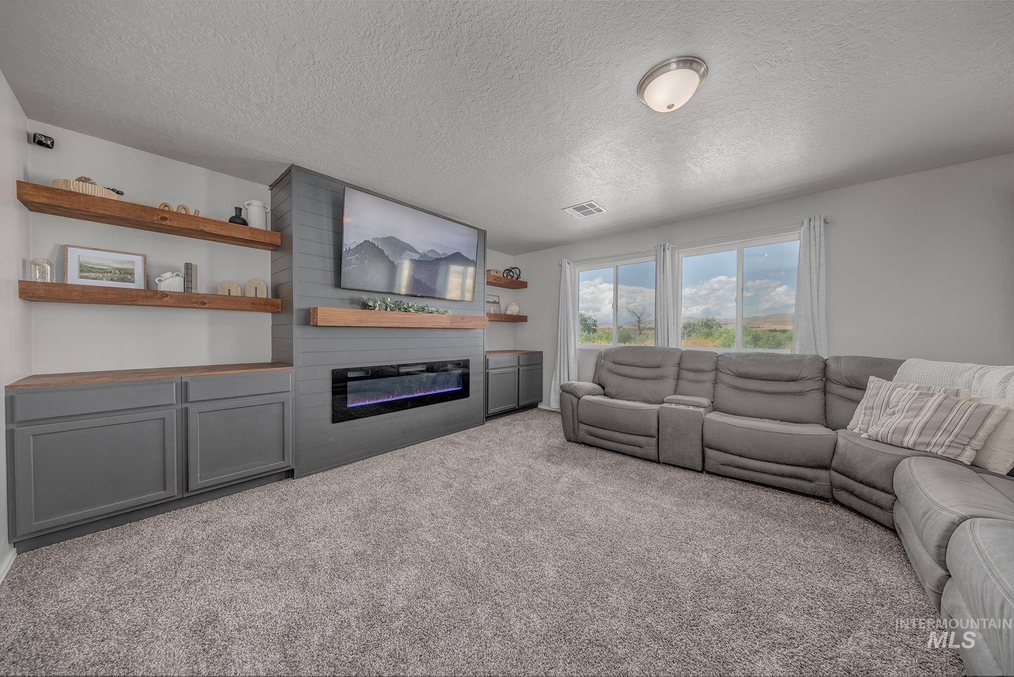 Living area featuring a textured carpet floor, a built-in media center with grey cabinetry and wood-finish countertops, floating wood shelves, and a linear electric fireplace