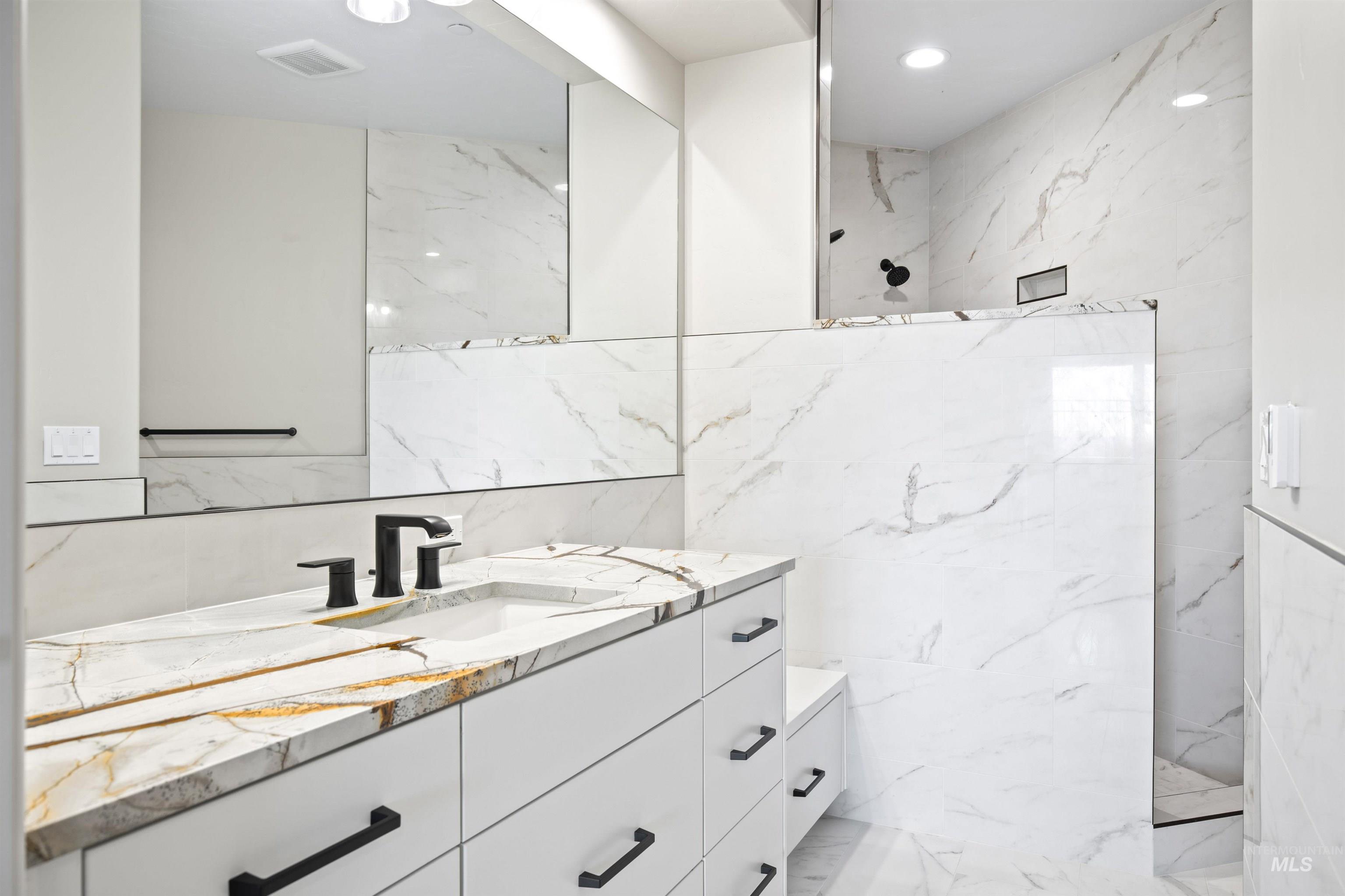 Bathroom featuring a white vanity with a gold-veined countertop, matte black hardware, integrated sink, large wall-mounted mirror, and a tiled walk-in shower