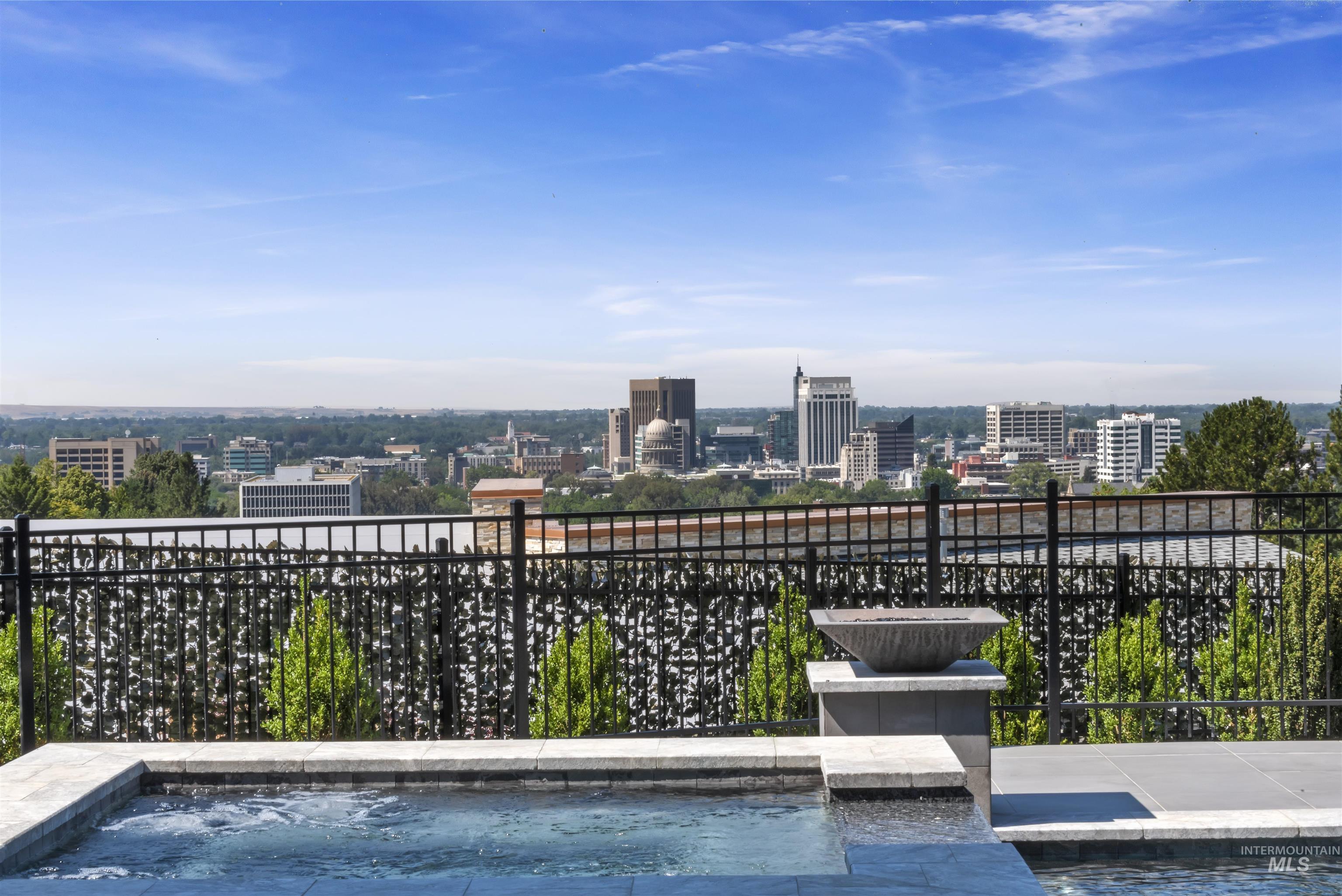 Outdoor spa with surrounding tile coping and pavers, featuring a black metal fence and an integrated fire bowl on a stone pedestal