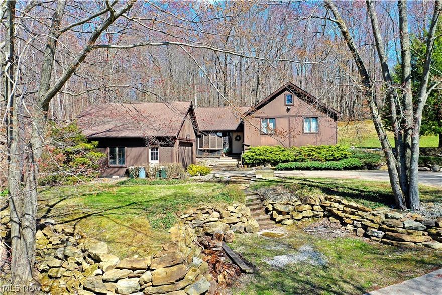 Uniqueness personified, this absolutely one-of-a-kind residence brings style, craftsmanship and a scenic, remote location with its expansive and tree lined lot! A winding concrete drive leads up the hill as select; hand placed stonework creates a series of walkways that outline the dark brown structure. Thick shrubs give way to a sloping hill that overlooks the relaxing rear sunroom and offset patio that overlooks a babbling nearby brook. Step inside to find a stone floor entry way filled with custom woodwork. The theme continues into the adjoining parlor that’s sat before the masterful living room. Exposed beams traverse each room to tie the far walls together as a brick fireplace sits below. A combined kitchen and dining area span the rear of the home with long running countertops that form bar top seating with its deep stained cabinetry. Eat-in dining offers easy sunroom access while windows wrap the corner. A half bath is located near garage access. Upstairs, three spacious bedroom