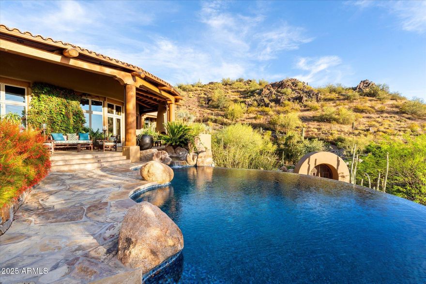 Hand-carved into the very mountain it rests upon, this extraordinary estate is more than a home--it is a livable work of art. Every element has been thoughtfully sculpted, from the hand-carved doors to the sweeping, curvilinear walls and ceilings, culminating in the striking wagon wheel ceiling that serves as a true architectural centerpiece. Rich in detail and artistry, the residence offers an immersive experience that continually reveals new layers of craftsmanship and design. Set within the exclusive and aptly named Lost Canyon, this is one of the most private properties in the area--yet just minutes from Scottsdale's private airport and the city's premier dining, shopping, and entertainment. Encompassing approximately 14 acres, the estate is surrounded by the breathtaking expanse of of the McDowell Sonoran Preserve, offering over 30,000 acres of protected desert landscape and more than 180 miles of scenic trails.
The outdoor living experience rivals that of a world-class resort--lush, immersive, and designed to transport you. Meandering pathways wind through the estate, following a tranquil stream and leading to a series of enchanting destinations. Towering natural surroundings and layered landscaping create a rare, Costa Rican-inspired atmosphere in the heart of the desert, where every turn feels like a secluded escape.
Throughout the grounds, ten fireplaces are thoughtfully placed, casting a warm glow that enhances the ambiance from intimate evenings to large-scale gatherings. From a secluded reading retreat perched at the upper reaches of the property, to a romantic grotto adorned with intricate stonework, every space invites exploration. Grassy play areas, swings, and a private golf chipping area further enhance the estate's park-like setting.
Entertaining is elevated to an art form with a rustic outdoor culinary pavilion, ideal for all-family cookouts and unforgettable evenings under the stars. Multiple seating areas, dramatic natural backdrops, and the soft flicker of firelight create an atmosphere that is both inviting and unforgettable--capturing the essence of a private luxury resort.
Inside, the home continues to impress with uncompromising quality and bespoke finishes. Venetian plaster walls, reclaimed oak flooring, and hand-carved mesquite doors speak to the level of craftsmanship found throughout. The kitchen is both functional and inspiring, with architectural detailing and panoramic views that make it a space you'll never want to leave.
A temperature-controlled wine tasting room showcases exquisite stonework, providing a setting as refined as the collection it houses. The primary office is equally remarkable, with custom millwork and thoughtful design that foster both creativity and focus.
This majestic mountain retreat is a rare offering--where art, architecture, and nature converge to create a home that lives like a private resort and feels as extraordinary as it looks.