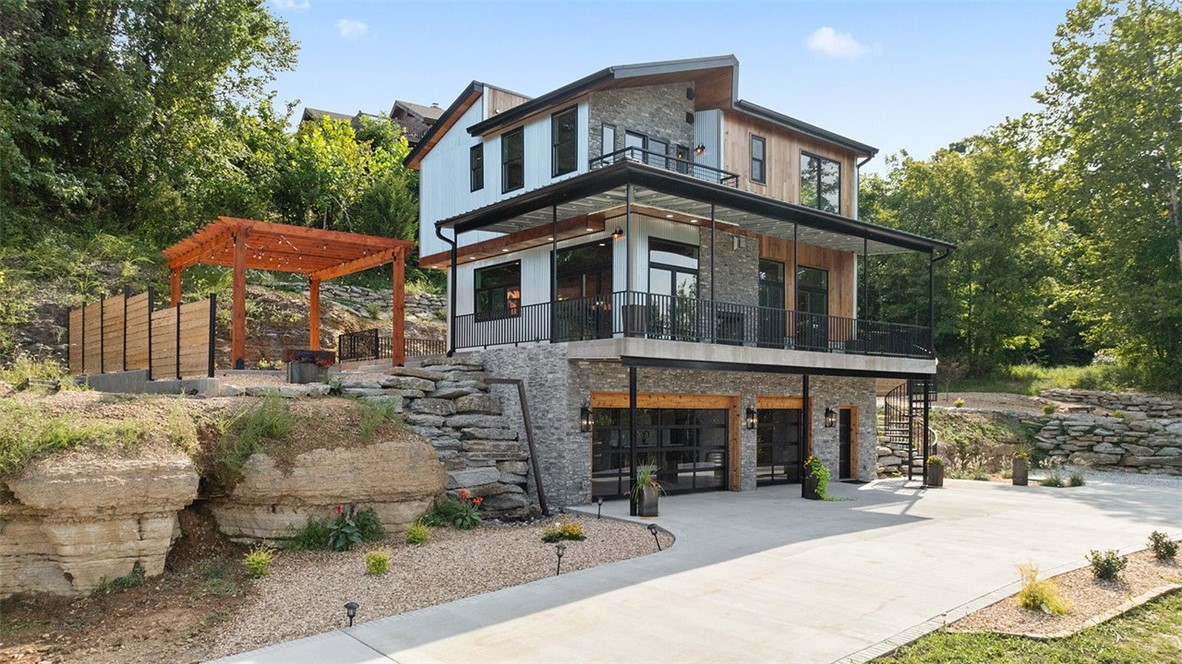 The Cliff House is an architectural short term rental masterpiece! This new construction home is perched just 10 minutes from Downtown Rogers, offering breathtaking lake views from every room. Designed for the best of modern living, the open-concept floor plan features soaring ceilings, custom tilework, a floating metal staircase, and a chef’s kitchen built for entertaining. A fireplace connects indoor and outdoor spaces. Step outside to enjoy a full-length deck, covered lounge, multiple patios, and a custom stone firepit, all positioned to capture the lake panorama. Amenities include a private wine and cigar room with a vault door and exposed cave, blending sophistication with originality. A geothermal system ensures efficient and sustainable comfort year-round. With unrivaled views, striking design, and an exceptional location near public boat launches, the Cliff House is a rare opportunity to own one of the most unique homes in Northwest Arkansas. The second parcel is also listed separately under MLS #1321493