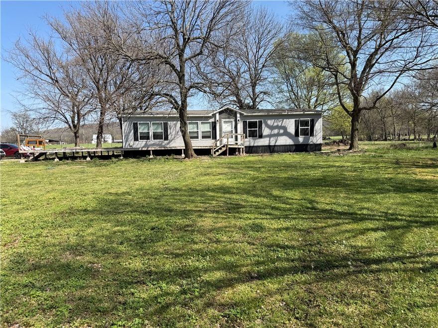 Located just 20 minutes west of the Wedington exit and 20 minutes south of Siloam Springs, this beautiful 5.8-acre property offers the perfect blend of country charm and convenience. The land is flat, open, fenced, and has paved road frontage—ideal for enjoying peaceful rural living with easy access to town. The 2022 mobile home features 1,512 sq ft with 3 bedrooms and 2 bathrooms, offering a warm and inviting layout. Inside, you’ll find LVP flooring, a spacious kitchen with island, furnished appliances, a walk-in pantry, cozy living room built-ins, and a private master suite with a walk-in closet. Step outside to the large deck and take in the quiet countryside, whether you’re sipping coffee in the morning or winding down at sunset.