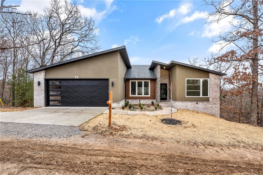 Move-in ready new construction by Elite Thomas Homes! This modern design features a sloped roofline with stucco, stone, and wood accents. Located on Bella Vista’s desirable east side, with the Taylor Homestead bike trail just behind the property. Large windows throughout the home capture beautiful wooded views from both levels. The main level offers an open living/kitchen/dining area with vaulted ceiling and beams, stacked stone fireplace, white oak flooring, and abundant natural light. The spacious primary suite includes a freestanding tub, dual vanity, tiled walk-in shower, and oversized walk-in closet. Bedroom 2 and a stylish guest bath complete the main floor. The partial walkout basement features a large media/game room, Bedroom 3, and a full bath, with premium upgraded carpet throughout. Two covered decks overlook the woods, giving you relaxing outdoor spaces to sip coffee, unwind at the end of the day, or entertain friends.