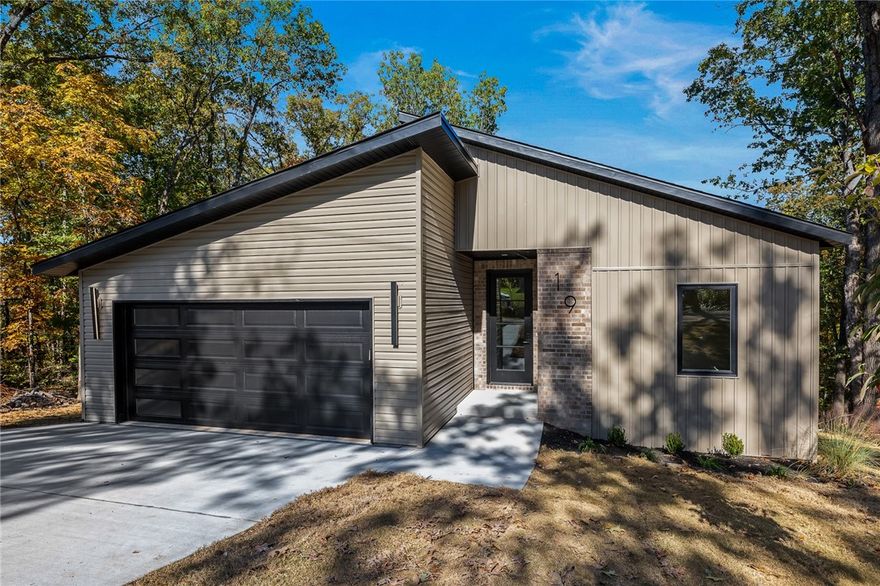 Located off a short gravel road on the east side of Bella Vista, this modern 1,900 sq ft home offers quick access to Pea Ridge and nearby bike and walking trails. Tucked into a quiet cul-de-sac and surrounded by mature trees, it provides exceptional privacy. The open-concept layout is filled with natural light and features luxury vinyl plank flooring, quartz countertops, a built-in drawer microwave, and propane hookup for a gas range. The living area includes an electric fireplace and opens to an oversized covered back porch overlooking a gently sloping yard and hardwoods. Additional highlights include sound wiring, pre-wired security, and a 1-year builder’s warranty.