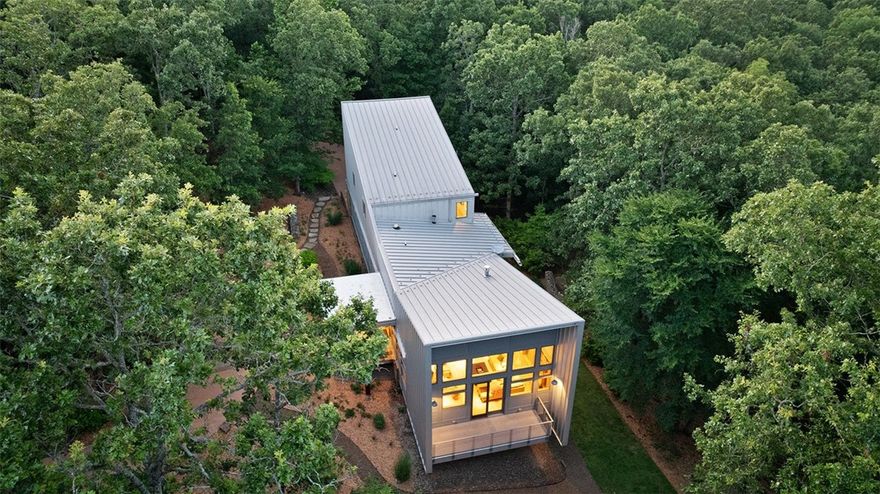 Introducing the Bowtie House—an architectural masterpiece nestled on 10 wooded acres in east Fayetteville. Designed by Tim de Noble, former Dean of Architecture at the University of Arkansas, this modern home blends seamlessly with the Ozark landscape. The unique “bowtie” roofline allows for vaulted ceilings, abundant natural light, and rainwater collection. The main level features the kitchen, living, dining, and a spacious primary suite—all universally designed for easy living. Floor-to-ceiling windows and an expansive porch create a seamless indoor-outdoor experience, with tree canopy views and seasonal glimpses of the Ozark Plateau. Interior finishes include maple floors, custom cabinetry, and cherry doors. The exterior is wrapped in Galvalume panels over stucco, wood, and native stone. Upper-level guest bedrooms offer privacy, while the lower level includes garage access and utility space. Peaceful, private, and thoughtfully designed—this home is truly one-of-a-kind.