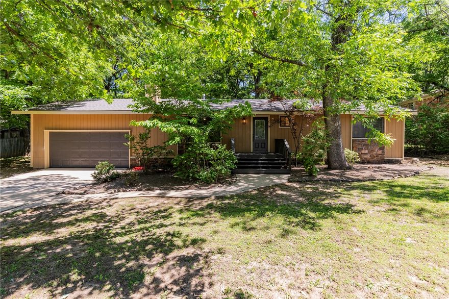 Tucked beneath a canopy of mature oaks just minutes from the University of Arkansas, this charming 3-bed, 2-bath ranch offers 1,590 sq ft of character and comfort in one of Fayetteville's most established neighborhoods. The stone-accented exterior, curved walkway, and lush natural landscaping create instant curb appeal with a wooded, private feel. Inside, hardwood floors anchor a warm living room featuring a stacked-stone fireplace and natural light pouring through oversized windows. The kitchen and dining area open to each other with tile floors, white cabinetry, center island, stainless appliances, and French doors leading directly to the back deck, perfect for entertaining flow. A standout primary bedroom features dramatic black custom built-in bookshelves, a moody designer touch that photographs beautifully. Step out back to a sprawling deck built around a mature oak tree, overlooking a fully fenced yard with room to play. Walk to class, Dickson St., Razorback games and all the energy of campus life.