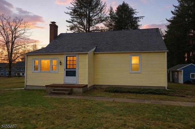 Yellow single-story house with warmly lit windows, a chimney, and a grassy front yard at dusk.