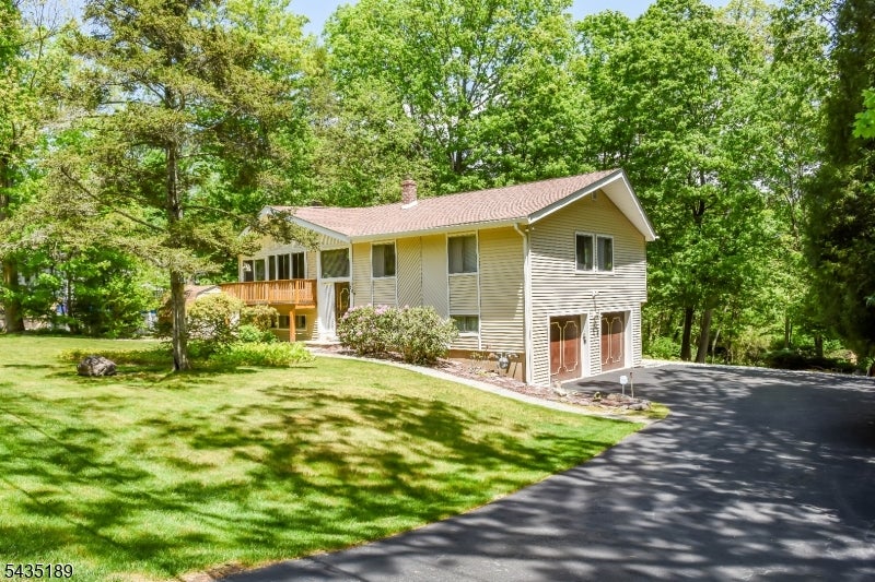 Beige two-story home with a raised deck, broad lawn, and paved driveway among mature trees.