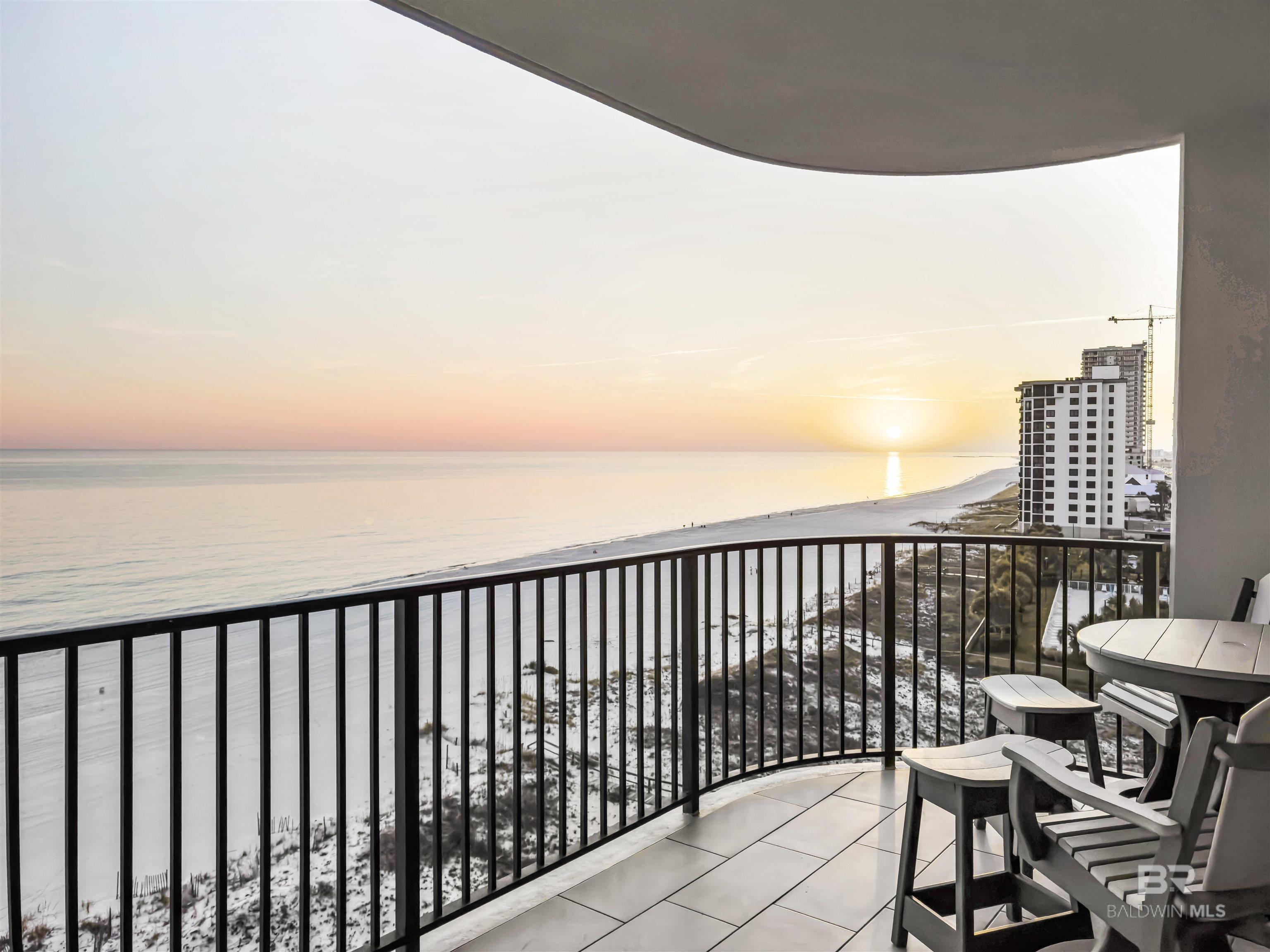 Furnished condo balcony overlooking the beach and Gulf at sunset, with a neighboring high-rise along the shore.