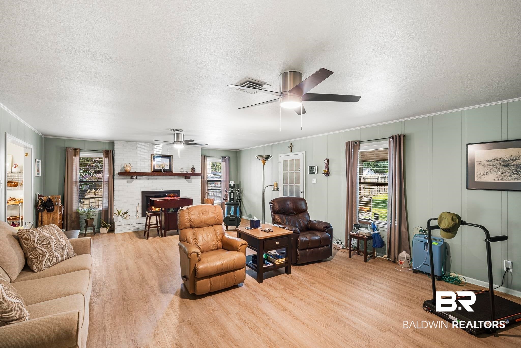 Spacious living area featuring wood-finish flooring, a white-painted brick fireplace with a wood mantle, and two ceiling fans