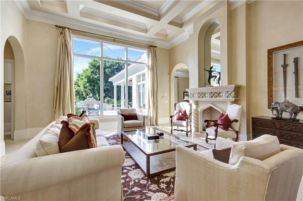 Elegant living room with coffered ceiling, large windows showcasing outdoor views, plush seating, and a decorative fireplace, highlighting luxury features of the Grey Oaks property in Naples, Florida.