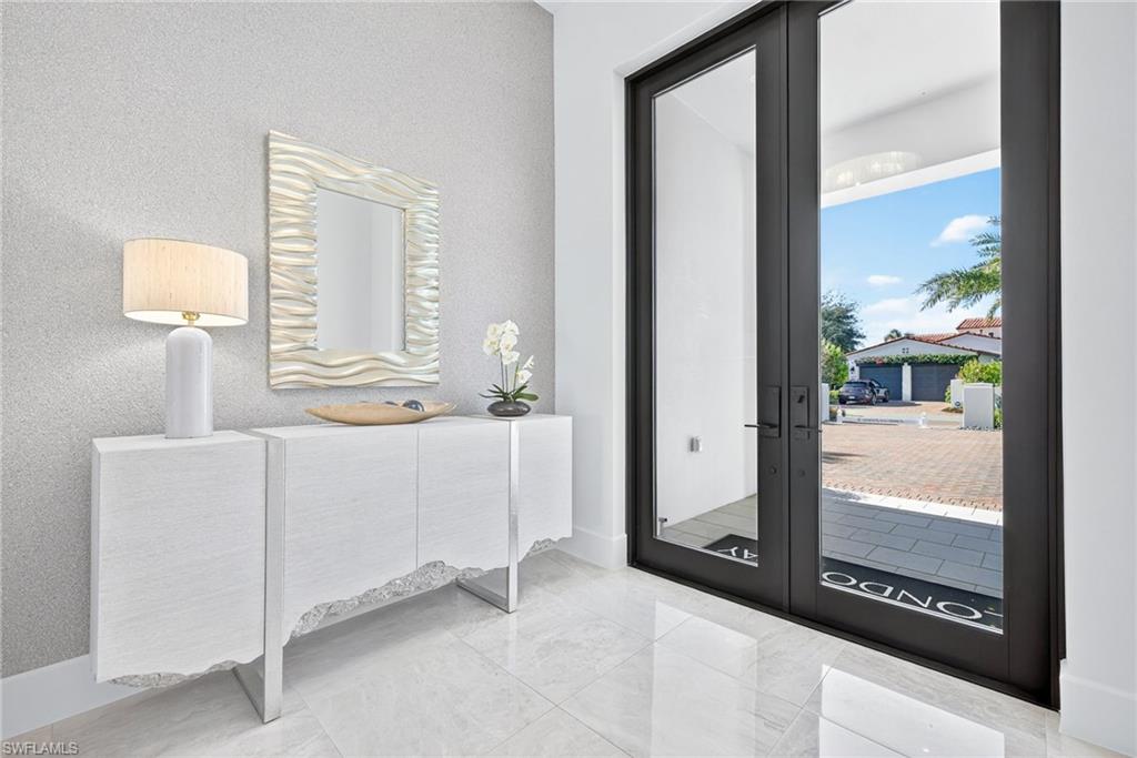Modern entryway with stylish furnishings, including a sleek console table and decorative lamp, showcasing a large mirror and fresh flowers, with large glass doors revealing a view of the outdoor area and driveway in a luxurious Naples, Florida home.
