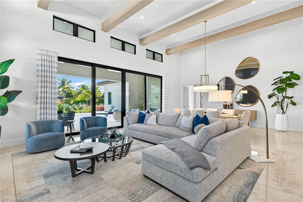 Modern living room interior featuring a spacious layout, large windows with views of tropical landscaping, stylish furniture including a gray sectional sofa and blue accent chairs, and elegant decor elements like a round coffee table and contemporary lighting fixtures, ideal for luxurious indoor-outdoor living in a new construction home in Lucarno, Naples, Florida.