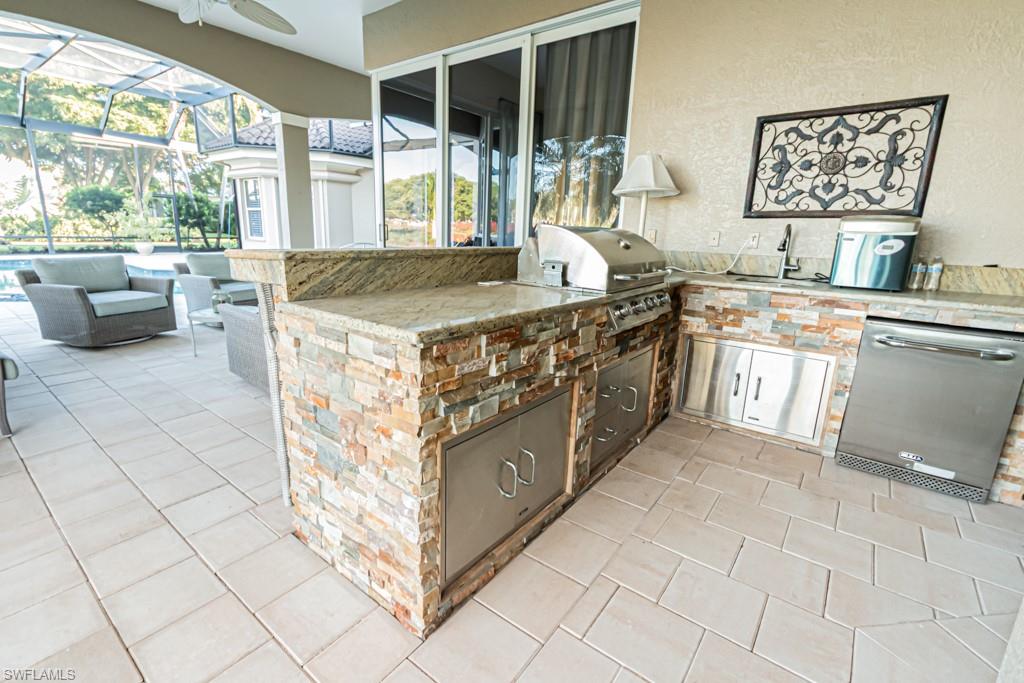 Outdoor kitchen with stone finishes, stainless steel grill, and sink, overlooking a pool area in a luxury home at Marsh Links, Naples, FL.