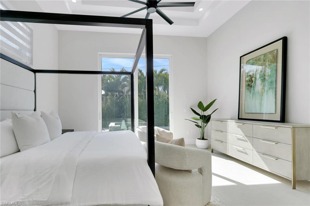 Modern bedroom with a four-poster bed, plush white bedding, and decorative pillows, featuring large windows with views of tropical landscaping, a stylish dresser, and a potted plant, highlighting luxury living in Naples Park.