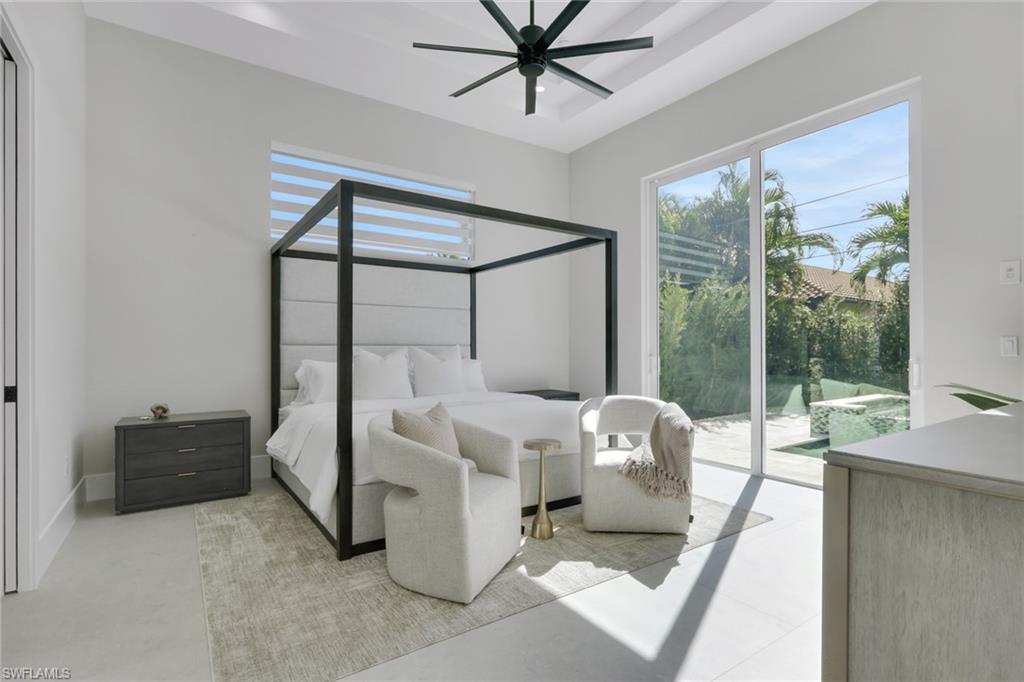 Modern bedroom featuring a four-poster bed, stylish armchair, nightstand, and large windows with views of tropical landscaping, emphasizing luxury and comfort in a newly constructed Naples Park home.