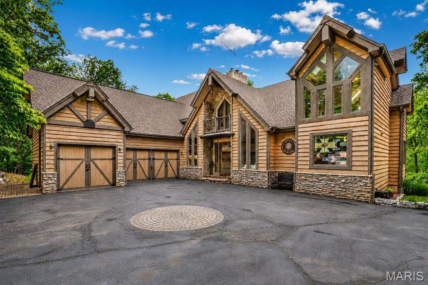 Tucked away on 4 breathtaking wooded acres, this extraordinary private estate offers the perfect blend of luxury, privacy, and natural beauty. Beyond the striking handcrafted gated entrance, a winding drive leads to a one-of-a-kind retreat surrounded by mature trees and serene views in every direction. Step inside the impressive foyer featuring handcrafted leaded glass and a stunning mosaic tile art wall. The foyer opens to a dramatic great room highlighted by soaring walls of glass, cathedral ceilings, lodge-inspired timber accents, and a magnificent two-sided wood-burning fireplace extending floor-to-ceiling. An impressive custom built-in entertainment center anchors the space while expansive windows showcase the surrounding wooded landscape. The adjoining luxury-appointed kitchen was thoughtfully designed for both entertaining and everyday living, featuring a massive 5x9 center island, handcrafted cabinetry, roll-out drawers and cabinets, and numerous custom finishes throughout. A second wood-burning fireplace creates warmth and ambiance, while custom doors lead to an expansive deck complete with a built-in gas grill. Adjacent to the kitchen are a formal dining room, spacious main-floor laundry room, and access to the oversized 3-car garage. The main floor also features the first of two luxurious primary suites complete with a private ensuite bath, walk-in closet, soaking tub, curbless walk-in shower, and direct access to a secluded private deck. Ascending to the second level, an open crosswalk overlooks the great room below and leads to an expansive office area, a charming sitting nook, and the second primary suite. This remarkable retreat features a dramatic full-wall library with ladder access to a third-level loft area. The ensuite bath includes a double vanity and another walk-in shower. As you descend to the lower level, you are greeted by one of the home’s most breathtaking architectural features — a spectacular curved custom staircase wrapped in soaring atrium windows. The lower level opens to an inviting family room with television viewing area, two additional bedrooms, and another full bath. Just beyond is an expansive recreation room with impressive 12-foot ceilings, ideal for entertaining, games, or additional gathering space. Additional features include 9-foot ceilings, 8-foot doors, casement windows, sliding barn doors, and a stainless steel outdoor wood-burning Hardy Stove capable of heating the entire home. The walkout lower level opens to an incredible outdoor living space featuring a large deck with a built-in fire feature incorporated into the stone columns. Overlooking the enchanting waterfall, tranquil creek, and private wooded grounds, the backyard also includes a rustic log bridge and a fire pit conversation area that creates a truly resort-like setting.