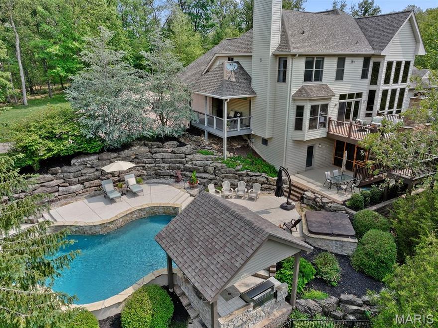 Discover your private sanctuary on over 3 wooded acres—an elegant custom retreat designed for privacy, comfort, and unforgettable entertaining. Enjoy your outdoor paradise featuring a saltwater pool with swim-up bar, spa, covered outdoor kitchen, full pool bath, and laundry with private entry. Relax on the composite deck overlooking the pool, complete with stone fireplace and built-in Green Egg grill. Step inside to a dramatic 2-story great room with soaring ceilings, a wall of windows, gas fireplace, and built-ins, flowing seamlessly into the gourmet kitchen with granite counters, large island, GE Monogram gas cooktop and hood, a stone backsplash, breakfast bar, and see-through wet bar. Adjacent is the hearth room with a floor-to-ceiling stone fireplace, offering access to a covered porch, perfect for relaxing year-round. The main-floor primary suite features a coffered ceiling, dual-sided fireplace, custom walk-in closet, and a spa-inspired bath with jetted tub, vaulted ceiling, walk-in shower, and double vanities. A breakfast room, coffered-ceiling dining room, and private office with built-ins complete the main level. Upstairs includes a loft overlooking the great room, with two spacious bedrooms and two full baths. The finished walk-out lower level offers 9' ceilings, abundant natural light, rec room with gas fireplace, bedroom, full bath, game room w/ bar, safe room w/ custom cabinets, workbench, and a wine/bourbon cellar. All just minutes from Missouri wine country.