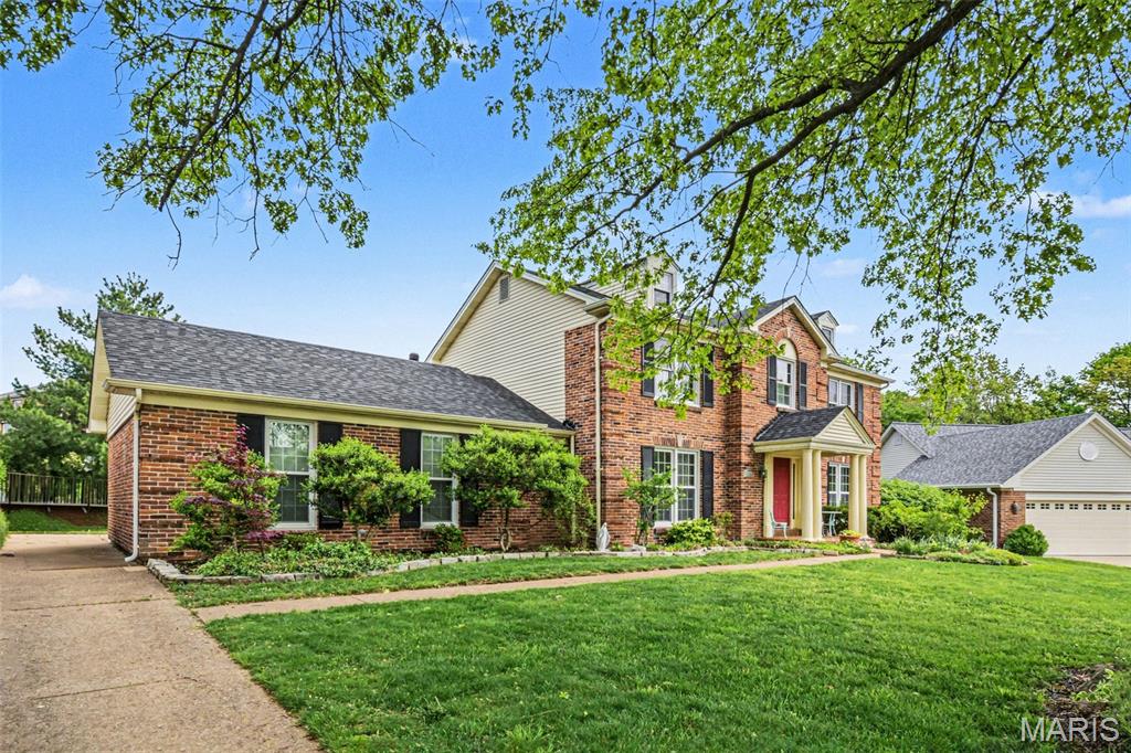 Traditional brick exterior with black shutters, mature landscaping, an attached garage, and a concrete driveway
