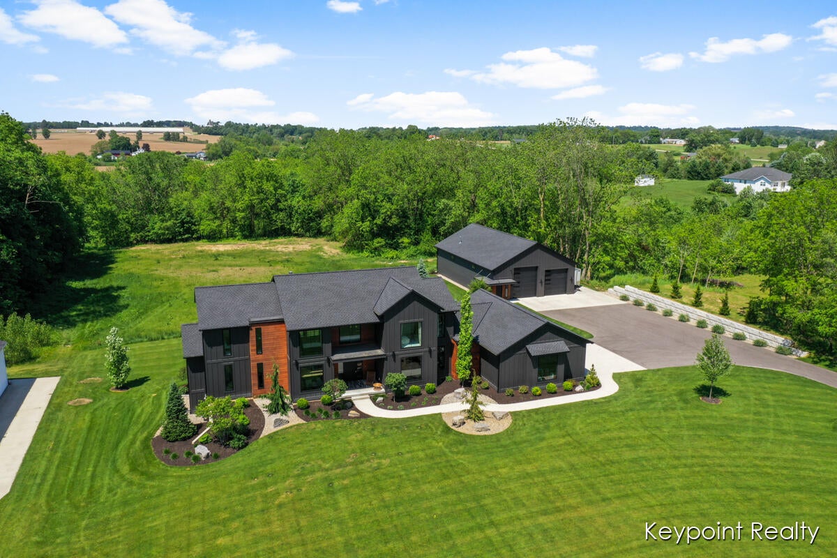 Aerial view of a modern dark-sided home with a detached garage, broad lawn, and wooded surroundings.