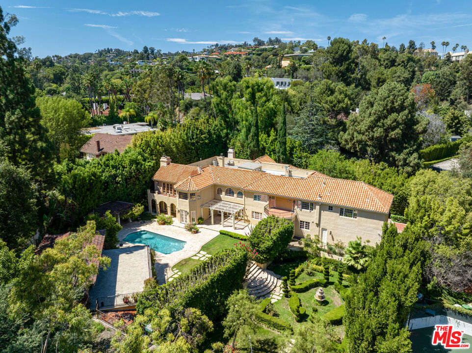 Nestled behind grand gates in one of Beverly Hills' most prestigious neighborhoods, lies a Mediterranean estate of unparalleled grandeur. The compound, with its manicured landscaping, offers the ultimate in privacy and seclusion, allowing for the full enjoyment of the custom pool, spa, sports court, and tended garden. Upon entering the double-height foyer, one is immediately struck by the opulence of the mahogany wood-paneled library, formal living and dining rooms, and the grandeur of the eat-in kitchen complete with full-size butler's pantry. The estate also boasts a gym and wine cellar, catering to the tastes of the most discerning individuals. The primary suite, a true sanctum of luxury, features a stunning chandelier, wood-burning fireplace, spectacular balcony, and dual exquisite custom bathrooms with walk-in closets. There are five additional en-suite bedrooms upstairs, along with an upstairs family room. Completing this idyllic compound is a detached guest house with a full kitchen, two bedrooms, one bathroom, and a rooftop cabana offering panoramic views of the estate. Host grand events in the expansive public spaces or retreat to the bedrooms for a moment of opulence, either way, 1055 Shadow Hill Way is a true embodiment of ethereal living