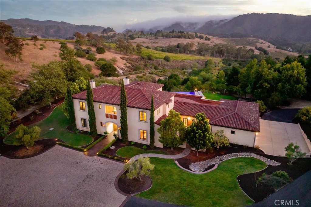 Tucked behind the gates of Bassi Ranch in the serene Avila Valley, this extraordinary property combines privacy, luxury, and timeless Tuscan-inspired architecture. Entering the gated community and turning down the private driveway, the setting captures your attention. Recessed below street level, the residence is hidden from view, offering privacy while framing sweeping views. The home sits on a 3-acre parcel with additional land for exclusive use of this property. The large 4 car garage is ideal for any vehicle enthusiast. A spacious motor courtyard welcomes guests with room for over 14 vehicles. The incredible custom floor plan provides the primary suite, a large office, a guest suite accessing the large, secluded courtyard and all living areas on the entry level to provide easy living. The house offers an extravagant entry with two story ceilings. The living room provides grand ceilings with wood beams and sprawling views of the valley and rolling hills. It also includes a wood burning fireplace and surround sound. The chef’s kitchen, designed for both function and beauty, has dual islands, double ovens, two sinks, two dishwashers, striking marble countertops, and space for everyone to cook and congregate together. The kitchen has beautiful picture windows overlooking the scenic Avila Valley and infinity pool. The kitchen and living room flow out into the back yard, where the heated infinity pool and spa overlook the horizon and surrounding valley views. The outdoor patio includes a built-in BBQ and sink. The partially covered patio area has built-in heaters. Mature landscaping ensures complete privacy. The primary suite is a sanctuary with an adjoining office, a large walk-in closet, and a spa-worthy bathroom. The shower features triple shower heads and a built-in bench. The primary bedroom leads to a private outdoor patio, adjacent to the pool and back yard. The spacious dining room is adjacent to the kitchen and opens to the private side patio. Upstairs provides a large den with sweeping views and two large storage rooms. It also has a deck to relax or greet your visitors. The two wings on the upper-level offer 4 total bedrooms and 2 guest bathrooms designed in a jack and jill orientation for ease and convenience. This estate truly embodies luxury living in Avila Valley’s unique microclimate while being moments away from San Luis Obispo and Avila Beach. It is designed for both relaxation and entertaining at the highest level.