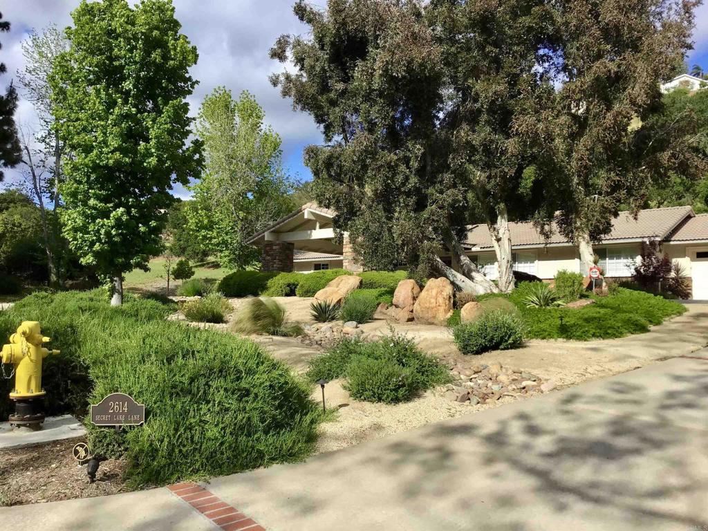 Serenity and privacy are effortlessly embodied within the highly coveted, and gated community, Secret Lake Lane in Fallbrook. Nestled on a scenic, tree-lined cul-de-sac, with a grand entrance that boasts a circular driveway, an elegant roofed porte cochère, a spacious three-car garage, and meticulously landscaped grounds. All accented by subtle tree lighting, which elevates the estate’s sophisticated design. This single-level residence spans 2,614 square feet and features four bedrooms, two and a half bathrooms, walk-in closets, a luxurious spa tub, and double vanities in both bathrooms. Sunlight gracefully filters through three skylights, highlighting the stunning vaulted wood ceilings, a striking floor-to-ceiling fireplace, natural hardwood floors, plantation shutters, and an additional fireplace in the living room. The gourmet kitchen showcases elegant sage and chocolate granite countertops, complemented by a matching wine bar, perfect for entertaining. Thoughtfully designed for versatility, privacy doors between the dining and living areas can be opened for social gatherings or closed for an intimate ambiance. The expansive 1.1-acre parcel is fenced and features meandering gravel paths weaving through nine ancient oak trees, illuminated by a sophisticated lighting system that highlights the outdoor beauty of the landscape. The outdoor living space boasts a charming flagstone fire pit with a pony wall, while privacy deco walls and a 16’x20’ Ramada with sun shades provide a cool, secluded retreat. For ultimate relaxation and entertaining, enjoy a seven-person spa, multiple patio seating areas, an outdoor fire pit, and an outdoor built-in sink. A separate side lot offers endless possibilities for a lush garden or personal creative endeavors. Located just minutes from Monserate Winery, Highway 76 and Golf Club of California, this remarkable estate offers a private sanctuary, blending natural beauty, sophisticated design, and exceptional privacy for an extraordinary lifestyle.