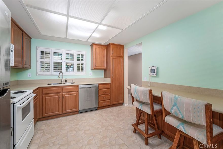 Welcome to this bright and inviting Villa Nueva model nestled in the desirable 55+ community of Laguna Woods Village. This spacious top-floor condominium offers serene treetop views and a comfortable floor plan designed for relaxed Southern California living. Entering the home, you’re greeted by durable tile-style flooring that flows into the kitchen and hallway. The kitchen features warm natural wood cabinetry, solid-surface countertops, and a full suite of appliances including a stainless-steel refrigerator, dishwasher, and sink paired with an all-white electric range and built-in microwave. A cozy breakfast bar offers additional seating and storage, while a window with plantation shutters fills the kitchen with natural light. 
The adjoining dining area features a ceiling fan and soft carpeting that extends into the large living room. Dual pane sliding glass doors open to a covered balcony, where you can enjoy tranquil views of the Aliso Viejo hills and Saddleback Mountain. The balcony’s elevated position provides privacy and quiet above the street, while the dual-pane windows throughout help maintain peace and comfort indoors. The living room is spacious and includes a custom-built entertainment center and shelving along one wall—perfect for displaying books, art, or collectibles. A heat pump ensures year-round comfort. The guest bedroom includes mirrored wardrobe doors, plantation shutters, and soft carpeting. The guest bathroom features a tub-shower combination with sliding glass doors, a large mirror, bright lighting, and ample vanity storage. The primary suite offers a relaxing retreat with plush carpeting, generous built-in storage, and a large bay-style window framed with plantation shutters. An accent mirrored wall adds depth and brightness to the room. The adjoining dressing area features a vanity with extra counter space, mirrored closet doors, and a private step-in shower with glass doors. This well-maintained home combines comfort, convenience, and the classic Laguna Woods lifestyle—complete with resort-style amenities including seven clubhouses, five pools, two golf courses, pickleball and tennis courts, fitness centers, equestrian trails, and over 250 clubs and social activities. Enjoy this peaceful retreat just minutes from the beaches, restaurants, and shops of Laguna Beach.