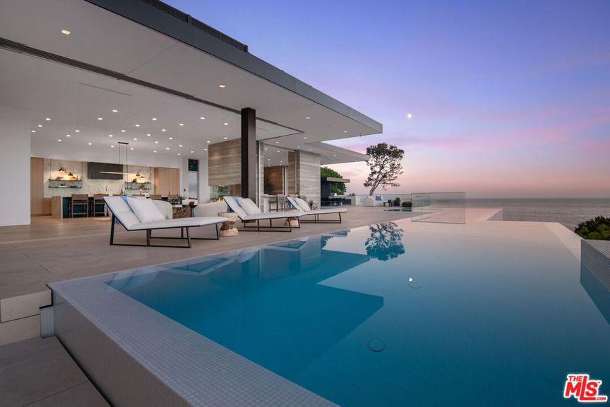New Construction | 2025 Mid-Century Modern Masterpiece with Breathtaking Ocean Views.Experience unparalleled luxury in this newly built, architecturally stunning mid-century modern home, perched on a bluff above Malibu Cove Colony. Nestled on the beach side of the Pacific Coast Highway, this estate boasts panoramic coastline views, offering an exclusive coastal lifestyle. Designed for effortless indoor-outdoor living, the main entrance showcases a stunning floor-to-rooftop water wall alongside a grand 12-foot glass pivot door. The open-concept great room boasts soaring 12-foot ceilings, a dramatic bay fireplace, and floor-to-ceiling glass sliders that seamlessly vanish into sophisticated stone pocket walls. Step onto the expansive wraparound deck, where a shallow wading/Baja area, Jacuzzi, and an impressive 93-foot lap pool come together to create the ultimate private retreat.This 7,356 sq. ft. residence offers five spacious bedrooms, six and a half bathrooms, a home office, gym (or 5th Bedroom), and a stylish lounge bar area with a media room in the lower level. Wake up to breathtaking ocean views from the main level, including the gourmet kitchen, where a massive 18-foot marble island and top-of-the-line appliances and wine fridge, set the stage for both intimate meals and grand entertaining.The primary suite, located on the main level, serves as a tranquil retreat with a spa-inspired bathroom, walk-in closet and studio/office. The expansive great room opens fully to the zero-edge infinity pool and outdoor entertainment space, where a built-in barbecue/outdoor kitchen, fire pit, and walk-up indoor-outdoor bar create a resort-like ambiance.Additional highlights include:* Floor-to-ceiling windows that bathe the interiors in natural light and ocean views* 12 x12-foot glass entry with stunning ocean views* 13-foot stone water wall at front entrance* Recessed lighting, skylights, and premium hardwood, stone, tile, and carpet finishes* Secure, gated entry with video intercom and an advanced security system* Laundry room with washer/dryer* Attached garage with guest parking. This Malibu masterpiece offers an unrivaled blend of elegance, comfort, and coastal sophistication. Don't miss your chance to call 26946 Pacific Coast Hwy home.