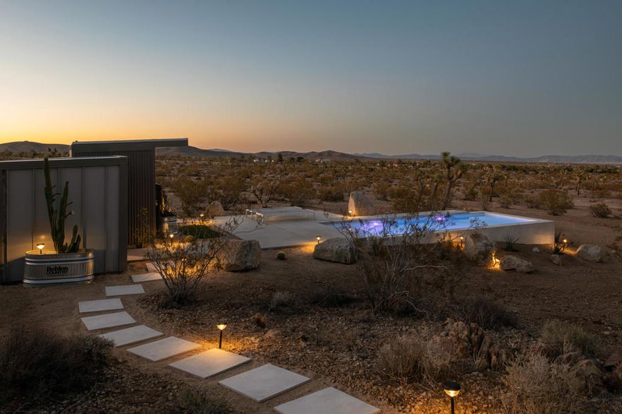 New 2024 architectural home in Yucca Mesa, set in a quiet hillside enclave with big, uninterrupted desert and mountain views. Built to a higher standard, the design is clean, calm, and filled with natural light. The oversized main living room anchors the home. Polished concrete floors and large sliding glass doors on both sides open completely to the landscape, creating true indoor outdoor living and framing the wide horizon. Two serene bedrooms sit at opposite ends for privacy. The main bath is beautifully finished with modern fixtures, natural textures, and soft light. Outside, the sculptural pool is the centerpiece. Set into the hillside and reaching over the desert to catch the sunrise, it invites long afternoons in the sun and quiet nights under a vast sky. A wood-lined sauna and a separate spa style bath add a rare wellness touch. Multiple terraces and a fire pit make gathering simple. Smart home controls manage lighting, sound, climate, and security. Surrounded by other design forward homes and minutes from Joshua Tree National Park, this property offers rare quality, privacy, and scale. Ideal as a primary residence, second home, or short-term rental investment.