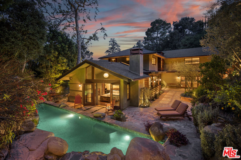 Exceptionally private and tucked at the end of a quiet cul-de-sac, this architectural retreat unfolds at the top of a long drive, surrounded by a breathtaking landscape of wild florals, mature trees, and layered landscaping. The setting feels like an enchanted garden, with a sense of peace that only deep nature and thoughtful design can provide. Floor-to-ceiling glass and large-scale windows create a seamless transition between indoors and out, allowing natural light and greenery to become a living part of the home's design. A stained-glass front entry greets with a kaleidoscope of color, leading to the grand living room boasting soaring ceilings and a dramatic sky-high gable window that creates ever-shifting light patterns as the day progresses. Wood beam accents and natural textures evoke the calm of a luxury mountain lodge, offering a true sense of escape.Nearby, the formal living room anchors the space with a wood-burning fireplace and opens through glass doors to a magical Wisteria-wrapped patio, offering treetop views and a quiet place to retreat. The dining room flows seamlessly into a warm, open kitchen centered around a handcrafted teak island and outfitted with Sub-Zero and Thermador appliances. The layout is designed for effortless connection, with the kitchen blending naturally into the home's main entertaining spaces.A glass lined gallery leads to the primary suite, offering a serene perch that looks out onto the lush gardens beyondan idyllic nook for reading or reflection. The primary bedroom is deeply tranquil, enveloped by the sound of cascading water from the nearby waterfall feature and the dappled shade of woodland-like greenery just beyond its doors. With its own fireplace, vaulted ceiling, and direct access to the backyard grounds, it functions as a private haven within the home. The spa-like en-suite bath includes dual vanities, a soaking tub with backyard views, separate shower and bidet, and a generous walk-in closet. Upstairs, a secondary suite offers sweeping views of the hillside and surrounding treetops, while two additional guest rooms, one with a balcony, share a large jack-and-jill bath. A lofted office above the living room serves as an inspiring perch, with treetop views, library shelving, and a soft soundtrack of birdsong that turns everyday tasks into a moment of calm.Outside, the home unfolds into a layered garden experience, with distinct spaces for dining, lounging, and quiet conversationevoking the timeless romance of Tuscany. A tall, ivy-draped wall frames a volcanic-stone fireplace with a custom heat-reflecting metal surround, anchoring the outdoor entertaining area with warmth and ambiance. Nearby, a built-in barbecue, alfresco dining table, and relaxed seating lounge complete the setting. The pool and spa are framed by natural rockwork and thoughtful landscaping, accompanied by a treehouse-inspired cabana with a full bathroom and sunbathing terraces tucked into private corners along the property. A lower-level garage offers two-car parking and connects discreetly beneath the home through a separate entry.Every detail from the tactile richness of the materials to the way light drifts through glass and foliagehas been composed to honor the landscape and evoke a sense of peace throughout the home. There's a transportive quality, as though it exists slightly outside of time: serene, enveloped, almost dreamlike.