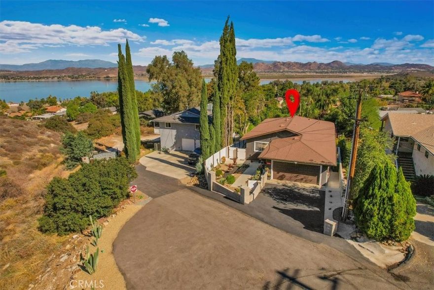 Tucked along a quiet incline in Lake Elsinore, this charming single-level home combines comfort, privacy, and beautiful outdoor living. A private walkway of dark wood leads to a tiled front porch and vibrant orange door, setting a welcoming tone. Inside, the home features cohesive ceilings with recessed lighting and wood-style flooring that extends throughout the main areas. The layout opens to a bright living and formal dining area, flowing into a spacious kitchen with an additional dining nook and sliding doors leading to the deck. A central hallway offers convenient access to the laundry area, two secondary bedrooms (one with garage access), a full bathroom, and the primary suite with an ensuite bathroom. Each bedroom includes ceiling fans and natural light. The true highlight of the home is its expansive, covered deck, crafted with rich dark wood, a ceiling fan, and serene views of the landscaped yard below. The backyard feels like a private retreat, featuring tiered spaces with stone and gravel pathways, lush greenery, and a spa tucked beneath the deck. The property also includes a two-car garage and a decorative stone perimeter fence, completing its inviting curb appeal.