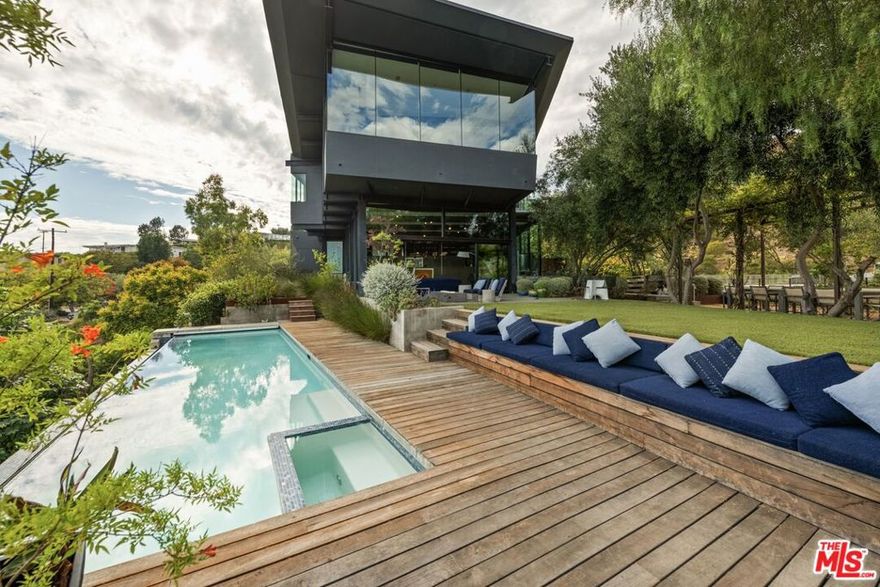 Designed by Hagy Belzberg, FAIA, OAA this striking residence showcases walls of glass framing sweeping canyon, city, and Santa Monica Bay views. A dramatic formal living room with floor to double-height glazing and sliding glass walls open to the outdoors to create seamless indoor-outdoor entertaining. Platforms that elevate the formal dining and kitchen allow all sides of this open plan to cascade from one space into the other. White oak floors and flamed finish salt stone throughout. A gourmet chef's kitchen with 11-foot island, Blue Star 60" range, Sub-Zero display refrigerator, dual sinks, two Miele dishwashers, and massive walnut open pantries. The primary suite is a private retreat with 300-degree mountain, city, and ocean views, a spacious lounge area, a built-in double-facing desk and office space, three walk-in closets - one with a sitting area and private terrace with frameless glass guardrails, and a spa-like bathroom with dual vanities and two separate shower entrances that meet in the middle, creating a dramatic glass-enclosed centerpiece. Free-standing two person tub with 180 degree views of deed-restricted open space. Additional bedrooms connect to a library loft with built-ins, complemented by a media room with custom workspace. Fourth bedroom is currently used as a gym. A generous driveway provides ample parking, complete with two electric car chargers for added convenience. Resort-style outdoor living includes an infinity pool and spa, built-in BBQ under a wisteria-covered pergola seating 16, and a desert garden with agaves, citrus, fire pit, and Palo Verde tree. A gated entry framed by hydrangeas and magnolias leads past a koi pond and bridge to a 14-foot glass pivot door, setting the stage for this architectural masterpiece.