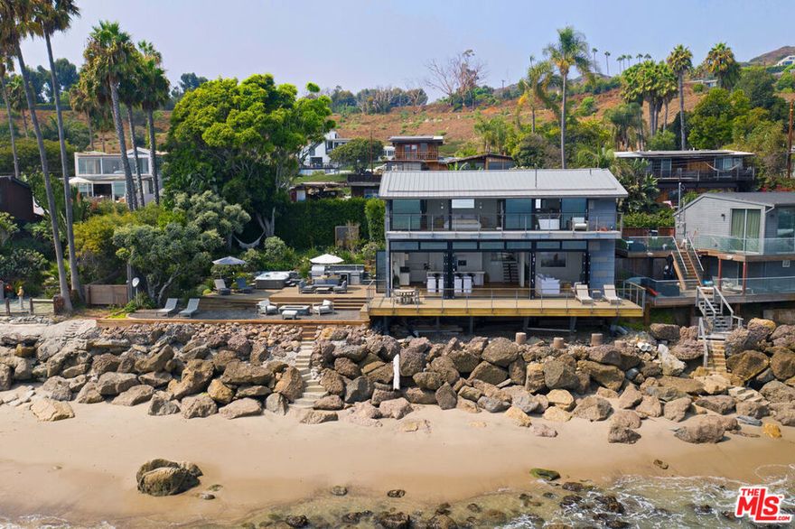 Updated contemporary on a double lot with rare 100 ocean frontage and a large landscaped lawn with hammock and a wood-and-glass sauna, perfect for lounging, dining, or working, and a detached guest room with a queen-sized bed and a 1/2 bathroom. A luminous entry hall to the surf break and non-stop ocean panorama, this stunning beach home with seamless indoor-outdoor flow, sensational views, and gorgeous open-plan design has everything you could possibly want for living the Malibu lifestyle. A private haven that is ideal for tranquil relaxation and sophisticated entertaining on a gorgeous cove with direct beach access, this special corner of paradise features wraparound decks, exceptionally high ceilings, skylights, enormous walls of glass facing onto the views, and beautiful wood floors. Step down from the entry to a sleek and sparkling great room with living, dining, and kitchen areas that open onto a beachfront deck through huge glass accordion doors, and to the entertainer's deck at the side of the house. The living room area has built-in shelving, a ceiling fan, an enormous flat screen, and a chic hanging fireplace in the corner. The open kitchen has a large island with bar seating, marble countertops, quality appliances, and temperature-controlled wine storage. There is one bedroom and a bath on the entry-level and two ocean-view, en-suite bedrooms, and a laundry room on the upper floor. The majestic master suite is enormous, with exposed beams, two ceiling fans, a fireplace, built-in shelves, a huge custom closet, a spa-style bath, and a wall of glass opening onto a full-width oceanfront balcony with ample room for seating and star-gazing. Accessible from the kitchen and from the lower-level deck, the entertainer's deck is furnished with a beautiful stone-clad outdoor kitchen and barbecue, a Jacuzzi brand spa, and ample seating and dining. Two steps down from the deck is a beachfront gravel area with additional seating, a fire pit, and steps leading to the beach. This amazing shoreline property also has air conditioning, automatic window shades, a speaker system, a carport, under-deck storage for surfboards and bikes, and breathtaking views over the mountains, coastline, ocean, and islands.