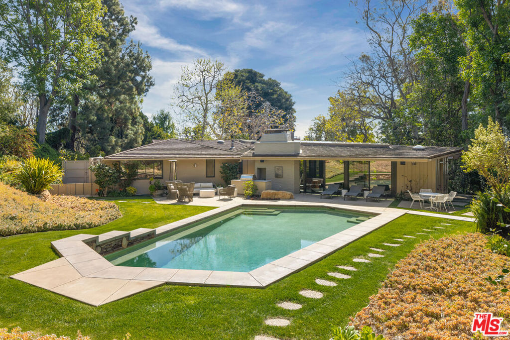 Tucked at the end of a private, gated drive in Brentwood's Mandeville Canyon, this Cliff May-style California Ranch home presents an extraordinary architectural statement amongst over an acre of lush park-like grounds. Initially built in 1951, this single-level modernist retreat invites a seamless connection between bright, open living spaces and the outdoors through walls of glass surrounding the entire backside of the home. Thoughtfully reimagined for contemporary living while maintaining the heritage of this iconic Mid Century style, the residence includes every luxury convenience sought by today's most discerning buyers. Enter to a sweeping open layout defined by grand, vaulted ceilings, exquisite original oak accents, and statement-making lighting including designer-selected pieces by Bocci, Artemide Logico, Dierk Van Keppel, French vintage finds, and a commissioned handblown "glass necklace". Ideal for grand entertaining, intimate gatherings, and everyday California living, the layout flows from a formal living room set around a stone fireplace, to a dining room with an original floor-to-ceiling built-in, and on to the family room and gourmet kitchen. The modernized kitchen offers functionality and style with a soothing blue-hued backsplash, abundant custom pantry storage, and professional-grade appliances -- SubZero refrigeration, Wolf double-oven range, dual Cove dishwashers, and a warming drawer. Filled with radiant natural light from skylights and Fleetwood glass doors that frame lush, garden views, the family room provides a dramatic focal point, outfitted with timeless pebble floors, a lava rock fireplace, and oak built-ins that lend depth and texture to the space. Down a thoughtfully lit gallery hallway that doubles as a personal exhibition space, find four of the home's five bedrooms, with the final bedroom on the opposite end. An expansive primary suite has a dedicated sitting room with access to a private patio, abundant custom cabinetry with solid walnut interiors, and an awe-inspiring spa bathroom clad in Calacatta Viola book-matched marble, Waterworks and Kallista fixtures, sculptural lighting, and boasting heated flooring, a soaking tub, and a dual, glass-enclosed steam shower. Flow outdoors to enjoy multiple areas to relax and entertain amidst the tranquility of the stunning canyon backdrop. Completely secluded for privacy and peace of mind, the more than 1-acre grounds are lush with mature landscaping, sprawling lawns, and vine-covered patios. Soak in the California sunshine poolside with the convenience of an outdoor shower and pool bath, dine alfresco with the built-in outdoor kitchen, and enjoy evenings under the stars from the hot tub. Other notable features include a laundry room with view of a custom mural, an attached 2-car garage with built-in storage, and a 10-car motor court set behind an automatic driveway gate. Exceedingly rare, this secluded Westridge Road residence is a true expression of the Brentwood lifestyle -- private, stylish, and moments from the celebrated dining, boutiques, and cultural institutions that define Los Angeles' coveted Westside.