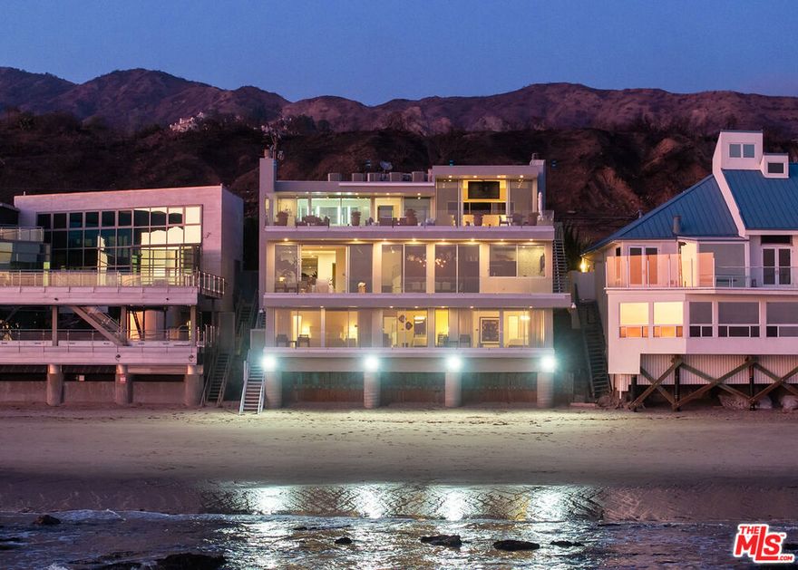 Beyond a sleek glass-and-metal front door, a luminous foyer welcomes with rich wood floors and a full-length mirror that amplifies the natural light. Straight ahead, walls of glass unveil panoramic ocean vistas that define the home's breathtaking setting. To the right, a refined powder room is appointed with stone tile floors, a stone vanity, and a shower with dual showerheads. An adjacent office offers soaring ceilings, recessed lighting, and warm wood floors, where sweeping ocean views enhance its distinguished atmosphere. The living room unfolds seamlessly from here, a showcase with a Crestron home system, floor-to-ceiling coastline views, and automatic shades for effortless comfort. Anchoring the space, a dramatic stone fireplace with matching hearth sets the tone for evenings by the sea. Beyond, a full-length deck provides an idyllic perch for ocean breezes and sunsets. The dining area flows into the living space, featuring custom cabinetry, a hidden bar with generous storage, a mini wine fridge, and an additional mini refrigerator. The open-plan design continues into the gourmet kitchen, where sliding glass doors frame the horizon. Stone countertops, dual sinks, a water filtration system, and a sliding service window complement premium appliances that include a Miele dishwasher, two KitchenAid refrigerator freezers, a KitchenAid microwave, and a stone-clad island with KitchenAid oven and Wolf electric range. Discreetly tucked away, the spacious garage provides double-car parking, integrated KitchenAid appliances, and ample storage. For peace of mind, the residence is equipped with a whole-house Generac generator and an advanced alarm system. On the lower level, a hallway lined with French doors reveals a laundry closet with stacked washer and dryer, plus a service room with extensive built-ins and a EuroCave wine fridge. With two full laundry areas and two washer and dryers in total, the home balances luxury with convenience. Each of the four bedrooms captures sweeping ocean views, a hallmark of the property's rare positioning. The first, located on this level, features wood floors, French doors, and sliding glass doors opening to an oceanfront deck. Its en-suite bath offers stone tile flooring, a vanity, and a steam shower. Further along, a staircase leads to a versatile basement level designed for a playroom or home theater, complete with built-ins, a cozy lounge, and an en-suite bath.Another guest suite mirrors the oceanfront charm with wood floors, deck access, and an en-suite bath featuring a vanity and shower-over-tub combination. The third bedroom impresses with floor-to-ceiling ocean views, a shared balcony, and an en-suite bath with stone finishes and a shower-over-tub. A fully outfitted gym with mirrored walls and professional flooring provides a private fitness retreat, while at the end of the hall, beach steps lead directly to the sand. Ascending to the top level, the primary suite awaits, an oceanfront sanctuary of elegance. Stone tile flooring, soaring ceilings, recessed lighting, and a grand fireplace define the room, while sliding glass doors open to a full-width deck complete with an outdoor dining area, lounge with firepit, and retractable roof. A ceiling-mounted television and integrated sound system add modern convenience.The spa-like primary bath features stone finishes, dual vanities, a deep jetted soaking tub positioned to frame the ocean, and sliding glass windows that open directly to the deck. A spacious steam shower with dual showerheads, a private water closet, and a large custom closet secured with a keypad lock complete the retreat.Blending architectural sophistication with coastal beauty, this Malibu Road estate is enhanced by rare amenities including a whole-house generator, dual refrigerator freezers, two full laundry areas, and four bedrooms with dramatic ocean views. An address of distinction, it offers the ultimate expression of luxury beachfront living.
