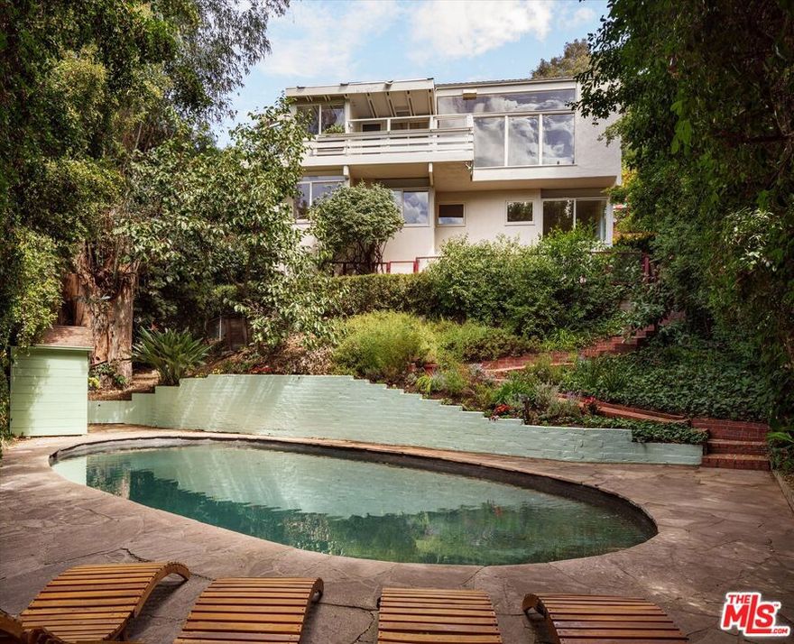 The Druckman Residence, circa 1941, by legendary architect Rudolph Schindler. Designed to integrate seamlessly with its surroundings, Schindler executed a residence that captures and communicates with nature. At the base of the garden sits a modernist swimming pool; an oasis in the middle of the city, more like a lake perfect in size and proportions. A previous listing of the property notes that the pool was designed by the great Richard Neutra. This aligns with Neutra's concept of giving occupants a visual and sensory connection to both water and to nature. Set back from the street behind a gate and a beautifully landscaped entryway, the house unfolds. Clerestory windows choreograph natural light throughout the day amid a palette of white plaster and original plywood surfaces. The main living area features a beamed ceiling, built-in cabinetry, and balcony, with large, south-facing windows that open to a grove of mature trees. Anchoring the room is Schindler's masterful fireplace, which continues to the exterior and an outdoor hearth, effectively creating twin living rooms, one interior and one al fresco on the property's large flat yard. The home offers quintessential modern Southern Californian indoor-outdoor flow, yet was decades ahead of its time. The kitchen frames treetop views from its original built-in table and breakfast nook, now complemented by refreshed surfaces and updated appliances. A second balcony opens from the kitchen to maximize light, air and access to nature.  An upstairs den completes this level, with verdant views and geometrical ceilings that echo Schindler's sculptural vision. The primary bedroom features large windows with canyon views, an expansive closet, and a newly updated primary bathroom. The three additional guest rooms each featuring canyon views, additional built-in, and bookcases along with backyard access. The Druckman Residence is both highly livable and historically significant, blending architectural integrity with privacy and convenience atop Outpost Estates, just moments from Hollywood, the famous Hollywood Bowl, and all that Los Angeles has to offer.