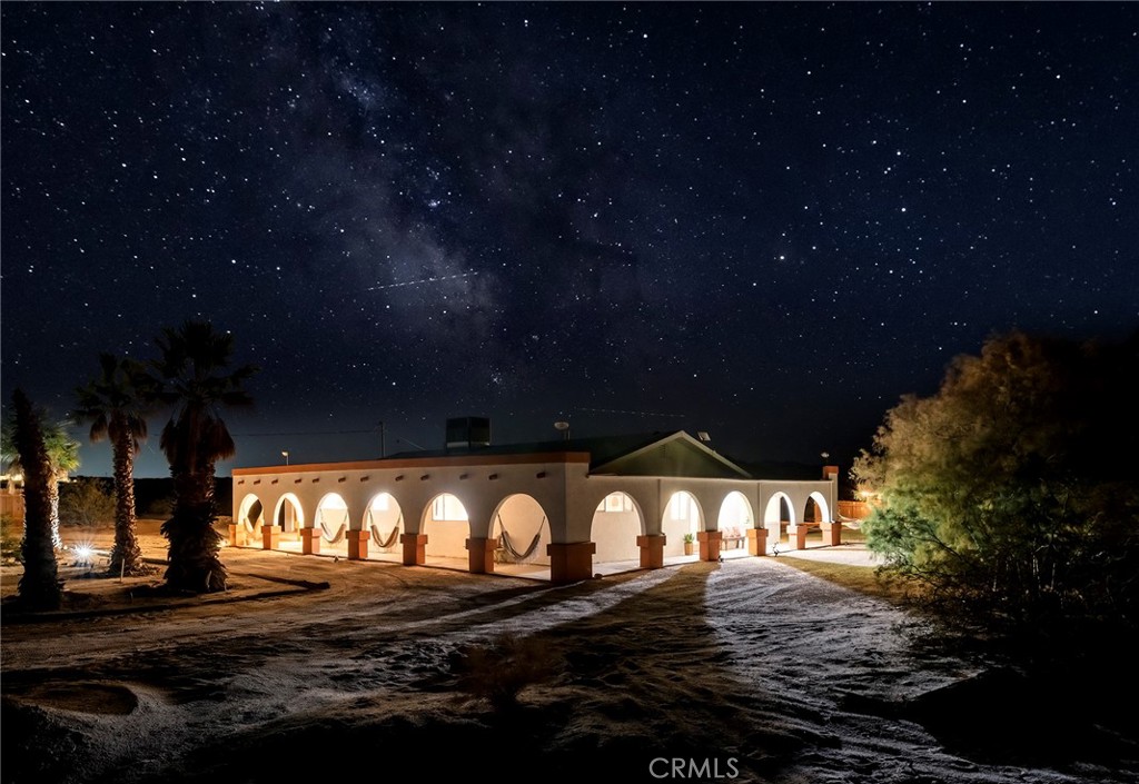 Welcome to Los Arcos, a striking Joshua Tree escape set in the heart of Wonder Valley—one of the most beautiful rural communities in the High Desert, known for its quiet solitude and some of the darkest skies in the Joshua Tree area. Here, stargazing is not just an occasional treat, but a nightly spectacle. This colonial Spanish ranch–inspired estate, with a playful 1950’s twist and its own Wild West saloon, rests on 5 private acres framed by mature trees and dramatic desert vistas. The property offers a rare combination of privacy, character, and convenience, located just 15 minutes from the eastern entrance of Joshua Tree National Park and downtown Twentynine Palms.
Currently operating as a successful short-term rental, Los Arcos is being sold fully furnished and turn-key, ready to continue its legacy as a beloved desert retreat—or to become your own private oasis in the High Desert.
Inside, the home is surprisingly spacious, with an open and inviting flow designed for comfort and entertaining. The updated kitchen blends retro charm with modern conveniences, while the bathrooms have been refreshed with thoughtful finishes that honor the home’s style. Multiple living areas provide room to gather or retreat in peace, capturing the easy rhythm of desert life.
Outdoors, Los Arcos invites you to linger. The grounds feature a spa, cowboy tub, outdoor shower, firepit, and hammocks for slow afternoons. Multiple shaded seating areas, framed by the property’s beautiful arches, offer cool respite from the sun. The original homestead cabin adds a layer of history and charm, while the attached two-car garage provides function and storage. And with a private well, you can enjoy desert living without water bills.
Whether you’re seeking a primary residence, a second home, or a proven short-term rental, Los Arcos offers a magical setting that’s as versatile as it is unforgettable.