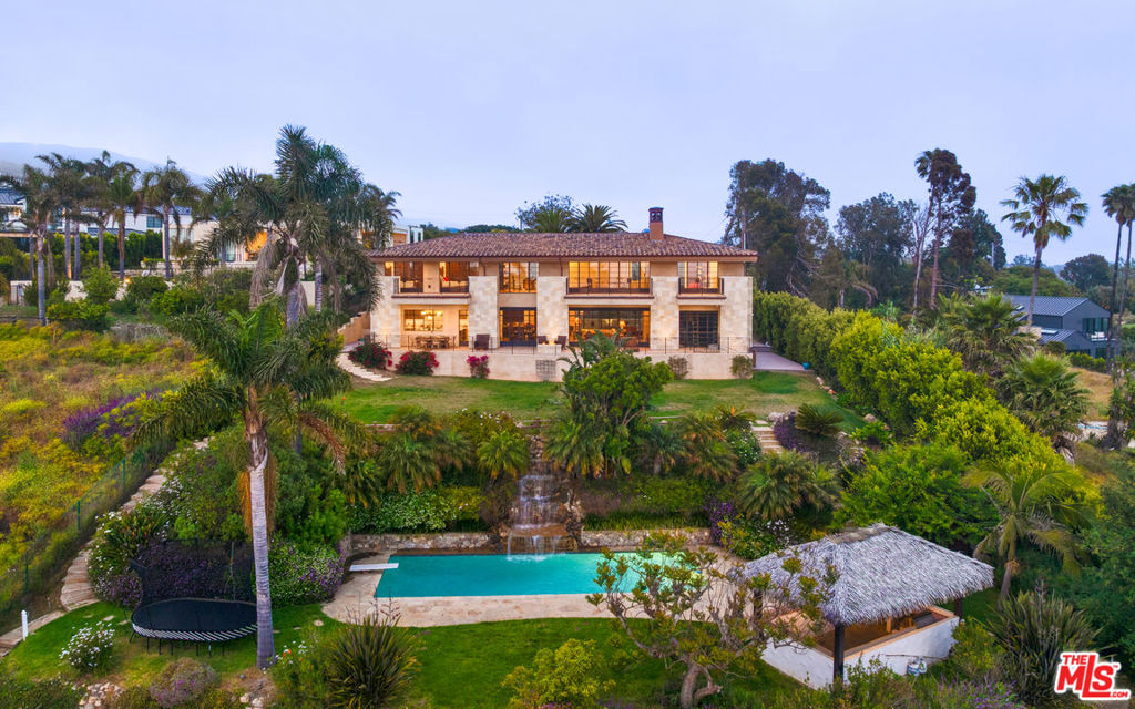 Tucked beyond a long, gated gravel driveway and nestled on nearly two private acres, this masterfully rebuilt estate offers an unmatched blend of refined craftsmanship and timeless design. Reimagined after 2018 with a commitment to non-toxic materials and natural finishes, the home is wrapped in imported stone sourced from an 850-year-old French chateau and features handcrafted doors from Arizona. Throughout the property, warm travertine and French oak floors meet artisan details like river bronze finishes and old-world character. Powered by a solar panel system and equipped with central heating and radiant warm board flooring, this residence is as sustainable as it is elegant. The front yard welcomes with manicured hedges, a peaceful fountain, and an expansive lawn. A grand wooden door opens to the main living area with soaring wood-beamed ceilings, a brick fireplace, and walls of glass-and-metal French doors framing panoramic ocean views. The open floor plan flows into the dining room, where double-height ceilings and a statement chandelier overlook the ocean-facing deck.The kitchen blends functionality and beauty with marble countertops, gold accents, and a large center island. Premium appliances include dual Cove dishwashers, Sub-Zero refrigeration, Wolf steam and microwave ovens, a six-burner cooktop with hood, and dual sinks one with a view of the ocean. A hidden bar with wine fridge complements the space, while a butler's pantry provides additional marble counter space, wine storage, and access to the backyard.The ocean-view deck spans the length of the home, with covered dining, a fire pit, and sweeping coastal views. Below, a flagstone terrace surrounds a sleek rectangular pool with a dramatic waterfall, framed by fruit trees and grassy lawns. The pool house includes built-in bar seating, a mini fridge, and barbecue station perfect for entertaining. Inside, an elevator connects all three levels. A hallway leads to a powder room with imported French tiles and a bonus room ideal for a projector theater. A three-car garage sits adjacent, while the lower level features a 2,000-bottle wine cellar with cigar storage, a second powder room, and a stone-tiled gym. The elevator's mechanical room and tankless heating system round out this level.Upstairs, the guest bedroom opens to a spacious ocean-view deck and features a steam shower, bidet, and generous closet. Additional bedrooms include covered balconies, built-ins, and a shared bath with double vanities. A dedicated laundry room is equipped with Electrolux machines and ample storage. An elevated office overlooks the living room, offering custom shelving, vaulted beamed ceilings, and unobstructed ocean views. The primary suite is a true sanctuary with high vaulted ceilings, a fireplace, French doors to a private balcony, and whitewater views. The ensuite bath includes a freestanding tub, double vanities, and a double-entry shower wrapped in rare stone. Two large dressing rooms complete the suite.Outside, enjoy citrus trees, a tennis court with lighting, lush gardens, hiking trails, and the calming sound of ocean waves. Offered fully furnished, this coastal retreat blends luxury living with mindful design.