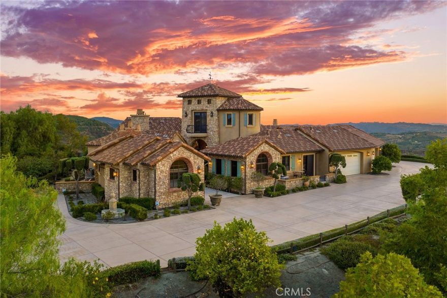 Welcome to Villa Girasole, a breathtaking Tuscan-inspired estate gracefully perched atop 15.6 acres in Temecula’s prestigious De Luz community. Evoking the romance of Italy’s hill towns, this architectural masterpiece blends old-world craftsmanship with modern luxury, surrounded by rolling vineyards, olive trees, fruit orchards, and hundreds of mature, producing avocado trees that blanket the hillsides in timeless beauty. Spanning approximately 6,588 square feet, this single-level residence was designed for both grand entertaining and intimate living. The thoughtfully curated floor plan features six bedrooms, seven bathrooms, a private executive office, a state-of-the-art home theater, and the signature Nexus tower. The tower boasts a stunning architectural focal point of the property, offering a curved staircase, arched windows, and panoramic balcony views of the Temecula Valley.

A separate, fully appointed casita with its own entrance, kitchen, living room, bedroom, and bath provides luxurious accommodations for guests or extended family.

At the heart of the home lies an extraordinary open-air piazza, an architectural centerpiece that connects every main living space. The saltwater pool and spa are sheltered within this elegant courtyard, framed by an outdoor fireplace, built-in BBQ, and a retractable projector for movie nights beneath the stars. This piazza was intentionally designed as the social core of the estate, offering a space equally suited for serene mornings or lively gatherings.
Inside, the attention to detail is unmatched. Vaulted wood-beamed ceilings, handcrafted brick archways, and intricate stonework create a sense of timeless permanence. The gourmet kitchen is a culinary showpiece, featuring dual center islands, a La Cornue range, built-in refrigerator, walk-in pantry, and an effortless flow into both casual and formal dining areas.

The primary suite serves as a tranquil retreat, complete with a cozy fireplace, spa-style bathroom, soaking tub, and views that stretch over the groves and valleys beyond.

Additional highlights include a six-car garage, four tankless water heaters, a private well with a 5,000-gallon storage tank, and thoughtfully placed olive and fruit trees enhancing the estate’s agricultural charm.
Villa Girasole is not just a home; it is a statement of elegance, privacy, and craftsmanship. A true sanctuary where architectural artistry meets the serene beauty of Temecula.