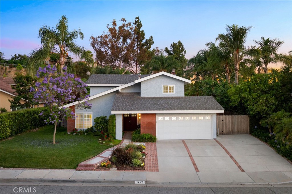 Situated on a peaceful street in Via Verde, this beautifully maintained two-story pool home blends thoughtful design with everyday livability. The manicured front yard showcases a mature Jacaranda tree in bloom, lush grass, colorful planters, and a brick-accented walkway that mirrors the driveway. Privacy hedges frame the home, creating a serene approach to the covered front porch and double entry doors. Inside, rich hardwood floors and soaring vaulted ceilings introduce the main living spaces. The living room features a large window framing the front yard, neutral carpeting, and a distinctive exposed post and beam detail. Adjacent, the formal dining room opens to the side yard through a sliding glass door and connects directly to the kitchen. The remodeled kitchen is finished with custom cabinetry, dovetail drawer construction, granite countertops, and a classic tiled backsplash. A peninsula with upper display cabinets, recessed lighting, and a sink with a backyard view add function and charm. The breakfast nook features a built-in buffet with upper cabinets and opens to the patio via another sliding door, ideal for indoor-outdoor living. The family room centers around a tiled fireplace flanked by a built-in entertainment unit, with space for a former wet bar that can easily be reimagined. A remodeled powder room with backyard access, plus direct garage entry, complete the main level. Upstairs, the primary suite includes view windows, dual mirrored closets, and a remodeled ensuite bath with coordinating cabinetry, granite surfaces, a custom tiled shower with frameless glass enclosure, and a dedicated vanity area. Three additional bedrooms, all with views, and a remodeled hall bathroom echoing the primary suite’s finishes complete the upper level. The backyard is a private retreat designed for entertaining and relaxation. A pebble-finished pool and spa with a slide, brick and concrete decking, a built-in barbecue, and a covered patio with retractable shade are surrounded by mature trees and lush landscaping, offering the feel of a secluded resort.