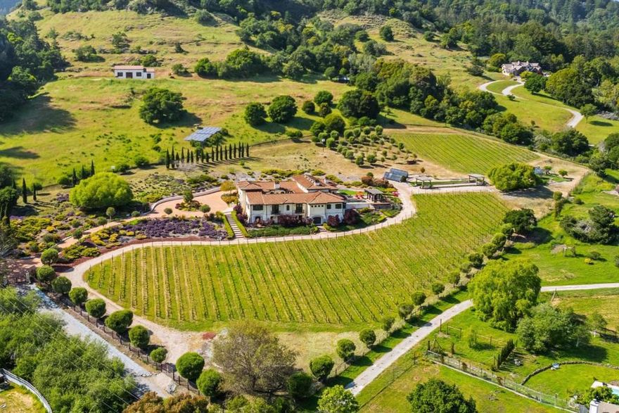 Nestled in a meadow & surrounded by over 21 acres of natural beauty, this magnificent gated Tuscan-inspired estate includes 1.5 acres of Pinot Noir grapes, offering a unique opportunity to own a private vineyard just 10 minutes from both Santa Cruz & Hwy 17 for easy access to Silicon Valley. At the heart of the single-level home lies a charming gated interior courtyard adorned with fruit trees, an outdoor fireplace, & a welcoming guest suite. The homes open layout, with abundant windows & French doors, invites seamless indoor-outdoor living. The spacious great room flows effortlessly into a chefs kitchen with other spaces including a formal dining room, family room & a pass-through bar opening to a built-in BBQ center beside the resort-style pool. The expansive grounds offer a bocce court, greenhouse, raised garden beds, a custom playground, a two-story treehouse with suspension bridge, pool & spa with designated bath, & multiple outdoor dining & lounge spaces. For automotive enthusiasts, an attached 4-car garage pairs with a separate 2-story barn/workshop featuring a car lift and room to accommodate up to 7 additional vehicles. The barn also includes a studio apartment & sits on a gently elevated section of the property, offering panoramic views & additional usable land.