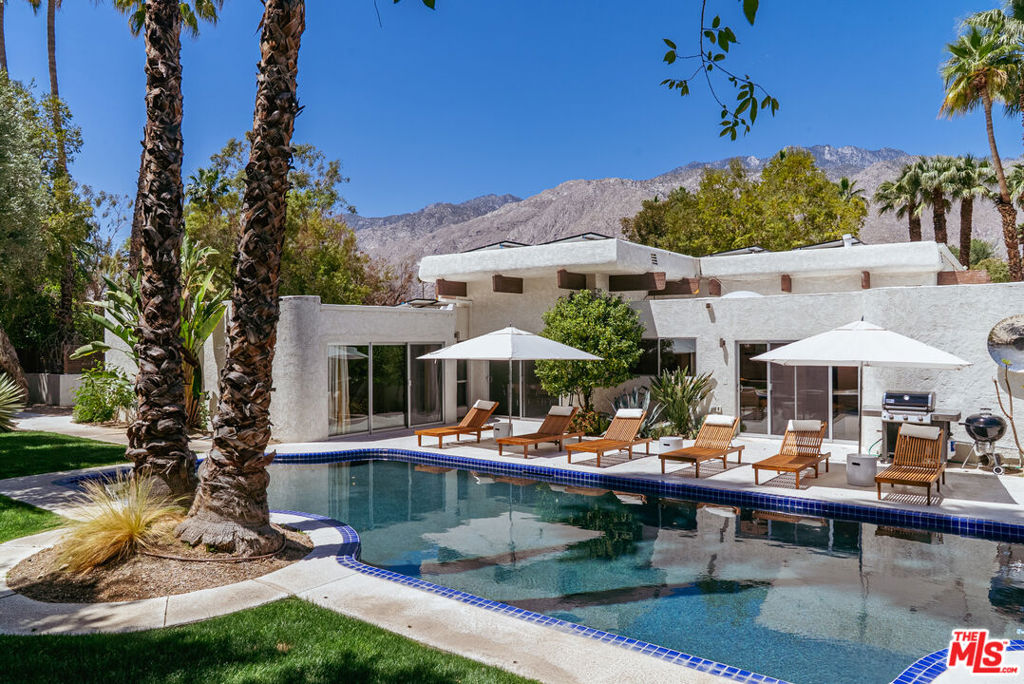 A stunning blend of modern sophistication and Palm Springs' architectural heritage, this estate is set in the coveted Movie Colony East, a neighborhood once home to Hollywood icons. Positioned on an expansive corner lot of nearly half an acre, it offers privacy and exclusivity, only attached to two neighboring properties. Located on the same street as Frank Sinatra's legendary Twin Palms Estate, it embodies a seamless fusion of history, luxury, and contemporary design. A dramatic sunken great room unfolds beneath soaring 15-foot coffered ceilings, anchored by a statement fireplace that enhances the grandeur of the space. Designed for effortless indoor-outdoor living, the layout features a formal dining area with lush poolside views, an open chef's kitchen outfitted with sleek Bertazzoni appliances, a natural gas range, a convection oven, and a breakfast nook perfect for morning rituals. The home offers three elegantly appointed en-suite bedrooms, along with an oversized attached casita that includes its own kitchenette and powder room, providing flexibility for guest accommodations or a private retreat. The primary suite is a true sanctuary, boasting a Finnish dry sauna, walk-in shower, soaking tub, and direct access to the pool, creating an experience of tranquility and indulgence. Step outside into a private oasis where native olive and palm trees, grape vines, and a citrus grove surround the pristine saltwater pool and spa, all set against breathtaking mountain views. Thoughtfully designed pergolas illuminated by bistro-style lighting create an inviting setting for alfresco dining, while a Weber char-broil 5-burner grill ensures effortless outdoor entertaining. Every detail is crafted for comfort and convenience, from the Ecobee smart system with dual-zone climate control to leased solar power for energy efficiency, an EV Level 2 plug for electric vehicle charging, and a spacious two-car garage with additional driveway parking. Ideally located just moments from downtown Palm Springs, golf courses, tennis clubs, museums, galleries, and fine dining, this estate offers both seclusion and accessibility. Beyond its architectural sophistication and luxurious amenities, the home is a study in effortless desert living, where mornings begin in sun-drenched tranquility and evenings unfold beneath a sky ablaze with stars. Whether hosting intimate gatherings by the pool, unwinding in the private sauna, or savoring alfresco meals framed by towering palms and majestic mountain vistas, every space is designed to evoke a sense of balance, beauty, and refinement. Exuding timeless desert luxury, this estate seamlessly blends architectural integrity with contemporary indulgence, offering a retreat that is unmistakably Palm Springs.