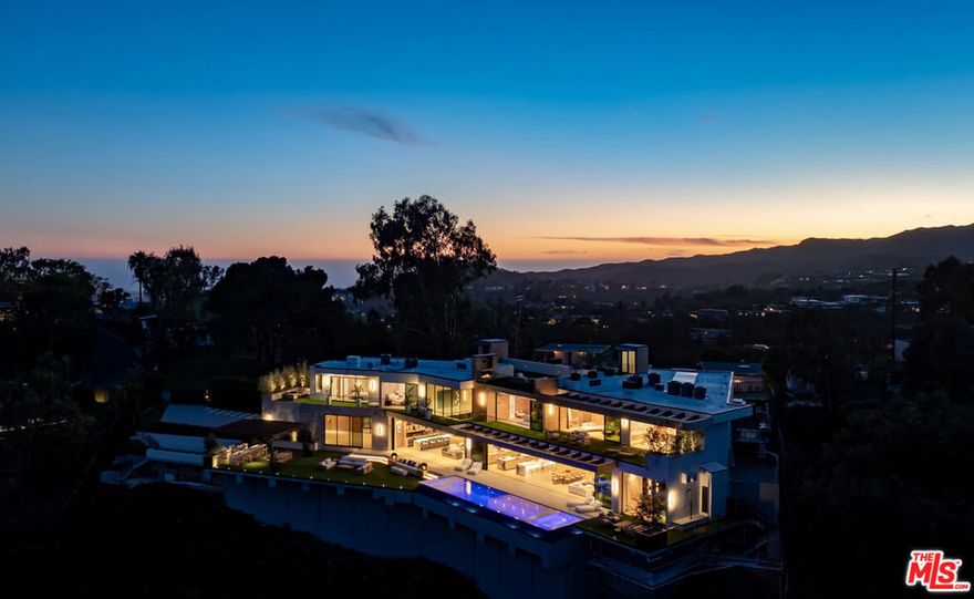 This newly constructed architectural estate, set on 1.2 sprawling acres, commands uninterrupted panoramic views of the city skyline and the iconic Getty Center. Designed with a mastery of form and function, every element has been thoughtfully composed to balance grandeur with intimacy. A herringbone stone motor court introduces the residence, leading to a glass-encased, air-conditioned four-car showroom. The entry is framed by a tranquil water feature and a lush living garden wall, setting the stage for the home's seamless integration of nature and design. Inside, elegant plastered walls and sky-high floor-to-ceiling glass create a gallery-like aesthetic, softened by warm travertine accents and European oak flooring.The soaring main living area is anchored by a dramatic double-height stone fireplace, floating terraces, and a striking olive tree centerpiece that blurs the boundary between indoors and out. A marble-clad bar with custom wood paneling offers an effortless transition to the pergola-covered outdoor kitchen and dining space, perfect for entertaining. The chef's kitchen is a showpiece, featuring a waterfall stone island, matchbook marble backsplash, a butler's pantry, a temperature-controlled wine showcase, and direct access to the laundry and staff quarters. Adjacent, the formal dining area, office/library, and one-of-a-kind sunken theater are all part of a flexible, open-concept layout that encourages both grand entertaining and intimate gatherings. Upstairs, the primary suite is a private retreat with a vast wraparound terrace, a spa-like ensuite encased in marble with a steam shower and soaking tub, and expansive walk-in closets. Five additional ensuite bedrooms, each with custom finishes and generous closets, surround an upper-level lounge and media room, designed for relaxation or more intimate entertaining. A dedicated wellness center offers a state-of-the-art gym, sauna, and steam room, while an elevator ensures seamless access throughout the home. Outside, vast terraces extend the living spaces, leading to an infinity-edge pool that appears to spill into the landscape beyond and overlooks otherworldly, head-on Getty Center vistas. Designed with solar capability, cutting-edge smart home integration, and a fully outfitted security system, this gated estate offers privacy, sophistication, and an architectural vision years in the making set against the breathtaking backdrop of Los Angeles.